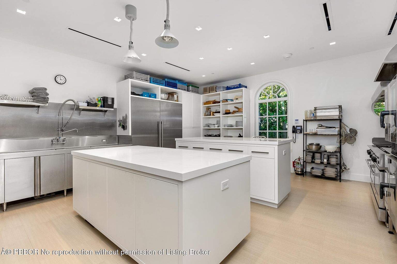 This is a spacious, professional-grade kitchen featuring stainless steel countertops and appliances, including a large refrigerator and a commercial-style sink. A large white island provides ample workspace, complemented by additional cabinetry and shelving for storage. The room is well-lit with recessed lighting and pendant fixtures, creating a clean and functional environment.