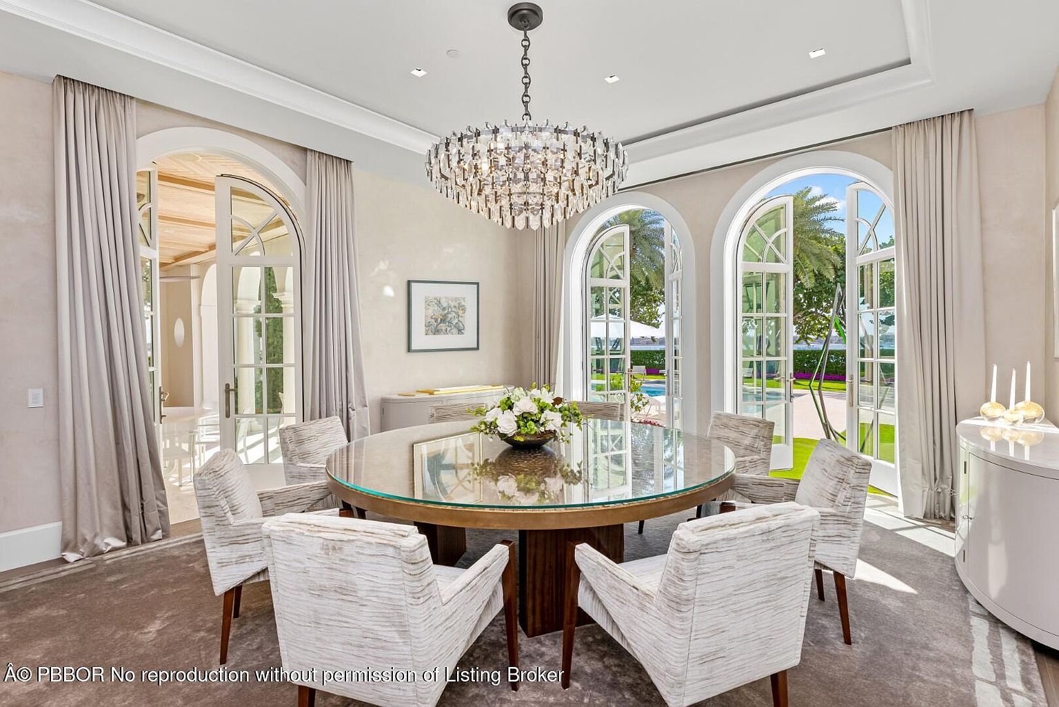This is an elegant dining room featuring a round glass-topped table surrounded by six upholstered chairs. A stunning crystal chandelier hangs above the table, and large arched windows with sheer curtains provide ample natural light and views of the exterior landscape. The room exudes a luxurious and sophisticated ambiance, perfect for formal dining.