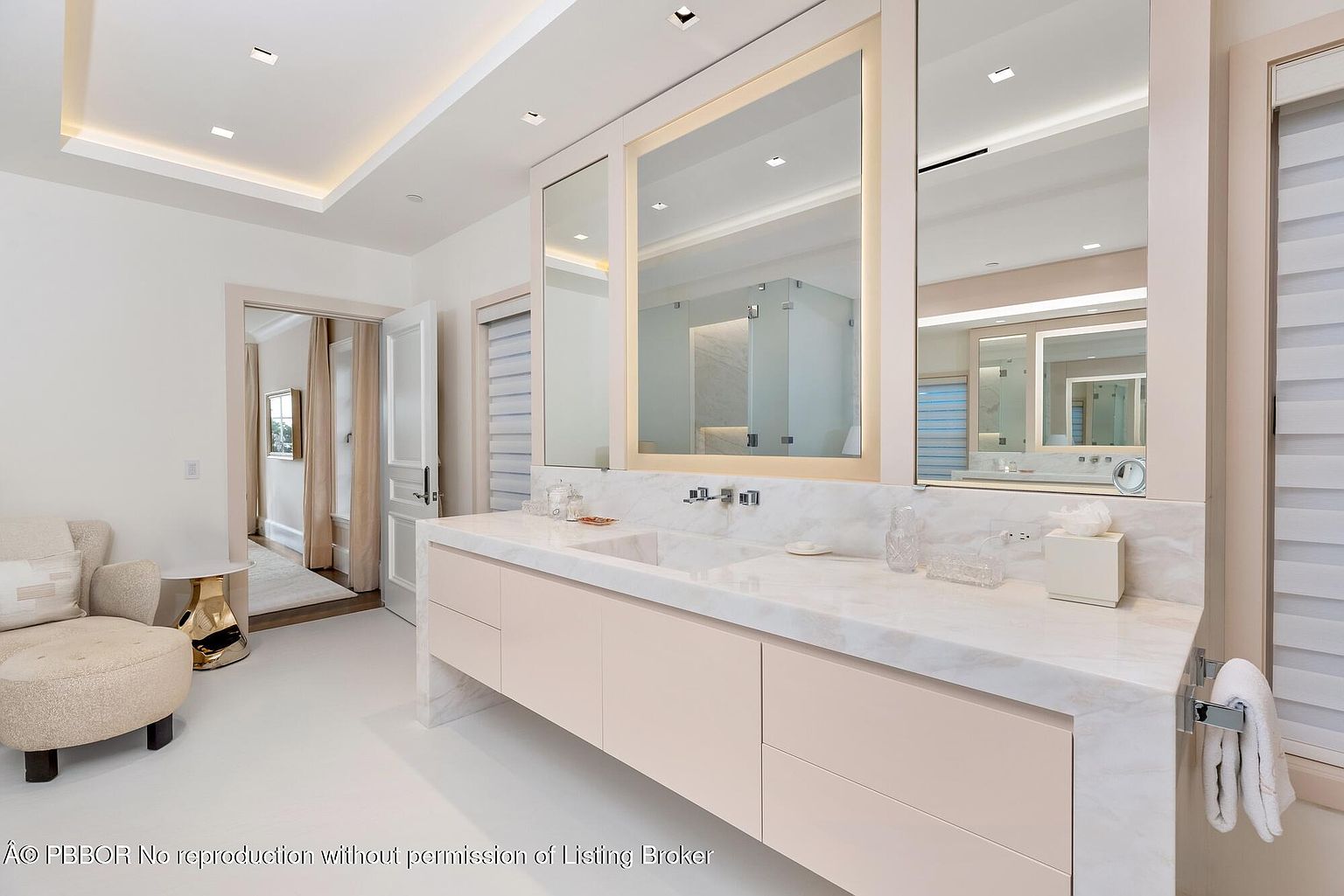 This is a bright and luxurious primary bathroom featuring a double vanity with a marble countertop and light pink cabinetry. Large, illuminated mirrors hang above the sinks, reflecting the clean, modern design. The room includes a seating area with a stylish chair and a glimpse into an adjacent room, creating a sense of spaciousness and elegance.