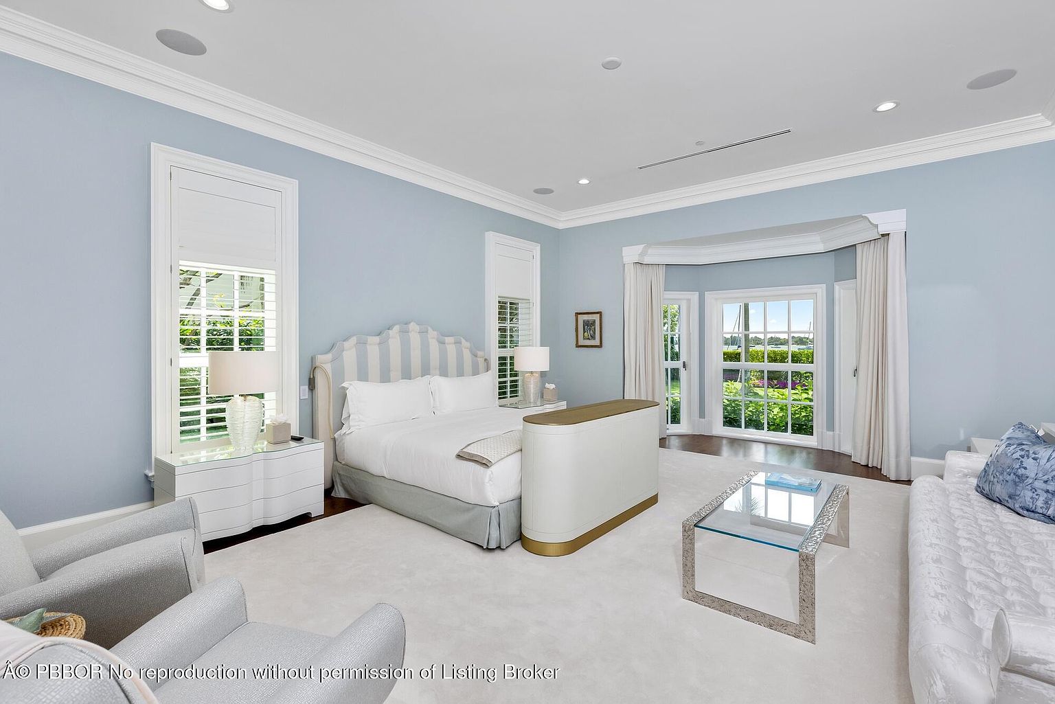 This is a primary bedroom featuring a light blue color scheme, a large bed with a striped headboard, and white bedding. The room has ample natural light from large windows with white shutters and curtains. A glass-topped coffee table sits on a white rug, and there is a seating area with upholstered chairs and a sofa, creating a serene and luxurious atmosphere.