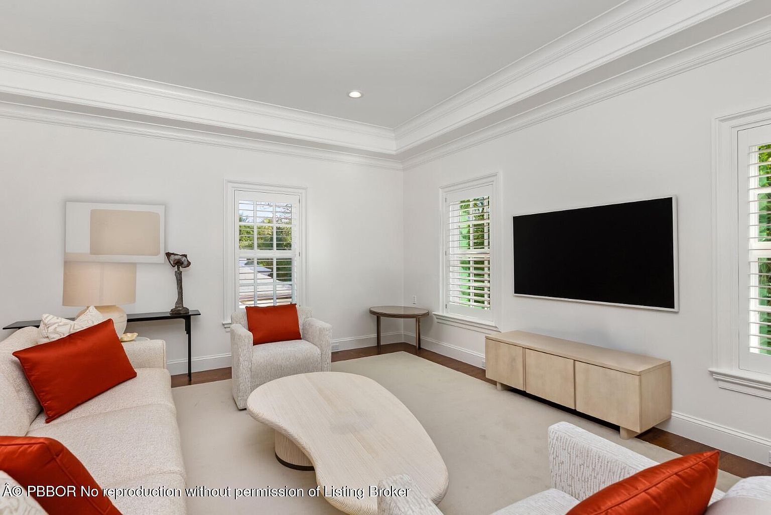 This is an interior shot of a living room featuring a neutral color palette with pops of orange in the pillows. The room includes a sofa, armchairs, a unique coffee table, and a media console beneath a wall-mounted television. Natural light streams in through shuttered windows, highlighting the clean lines and modern design.
