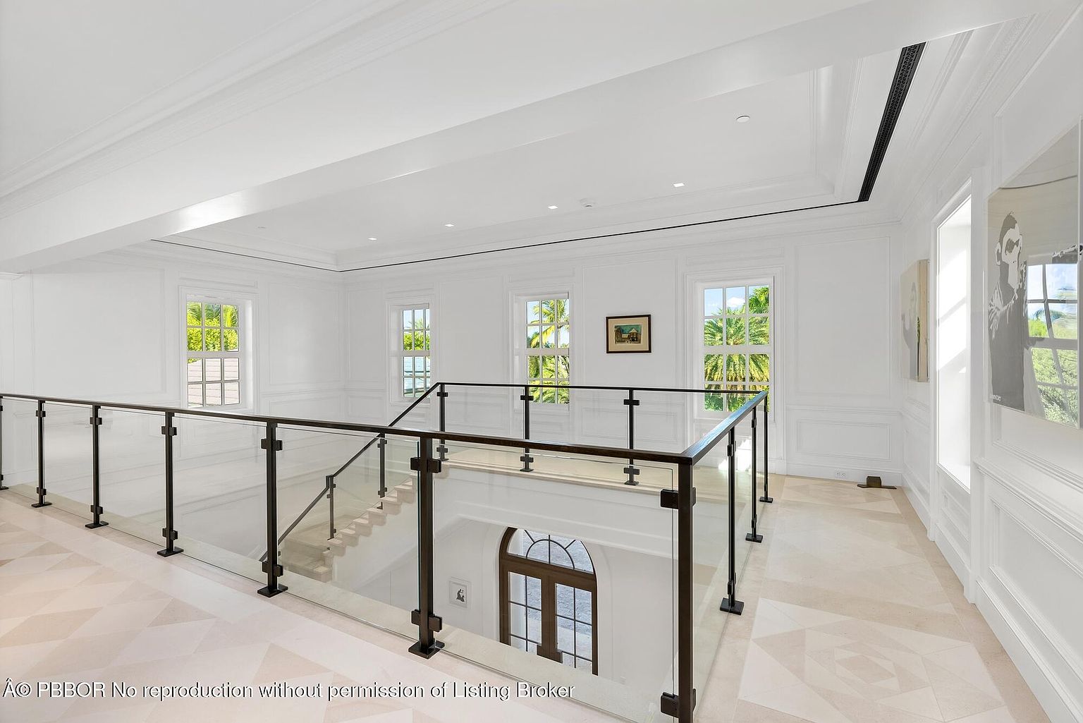 This interior shot showcases a bright and elegant hallway with a staircase. The hallway features a glass-paneled railing with dark metal supports, light-colored patterned tile flooring, and white walls with decorative molding. Several windows allow natural light to flood the space, highlighting the clean and modern design.