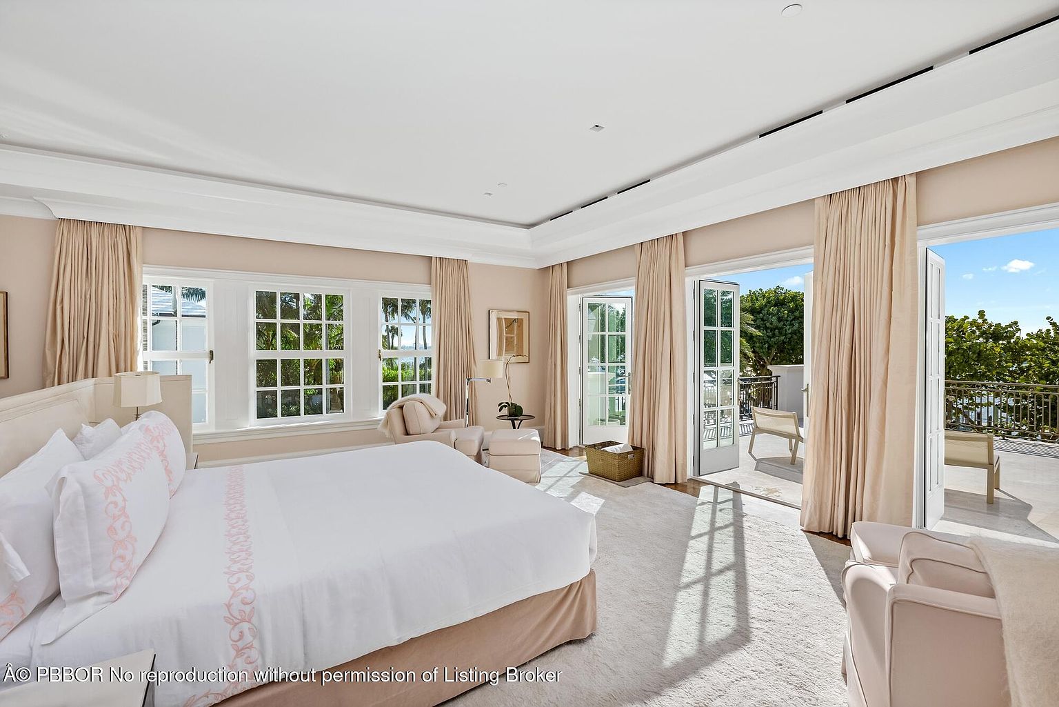 This is a bright and airy primary bedroom featuring a large bed with white linens and pink accents, multiple windows offering natural light, and access to a balcony through French doors. The room is decorated in a neutral color palette with light-colored curtains and carpet, creating a serene and luxurious atmosphere. The balcony offers an outdoor extension of the living space.