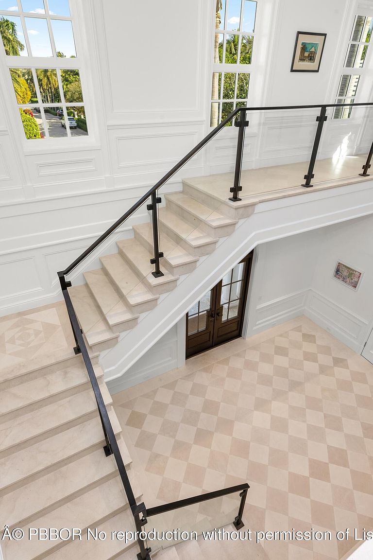 This interior shot showcases a grand staircase with light-colored stone steps and a modern black railing with glass panels. The hallway features a patterned tile floor and white walls with decorative molding, creating an elegant and bright atmosphere. A set of double doors is visible beneath the staircase.