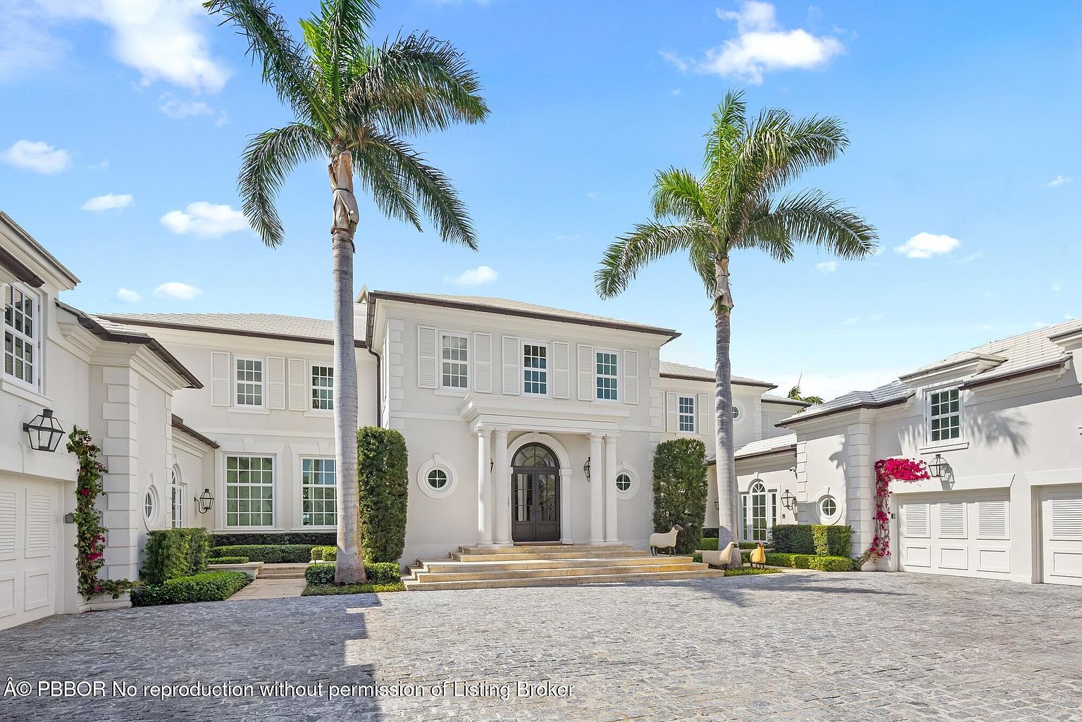 This is a front view of a grand, two-story white mansion with symmetrical design elements. The entrance features a covered portico with columns, leading to a dark-colored front door. Two tall palm trees frame the house, and manicured greenery and decorative sheep statues add to the curb appeal. The driveway is paved with cobblestone, enhancing the luxurious feel of the property.