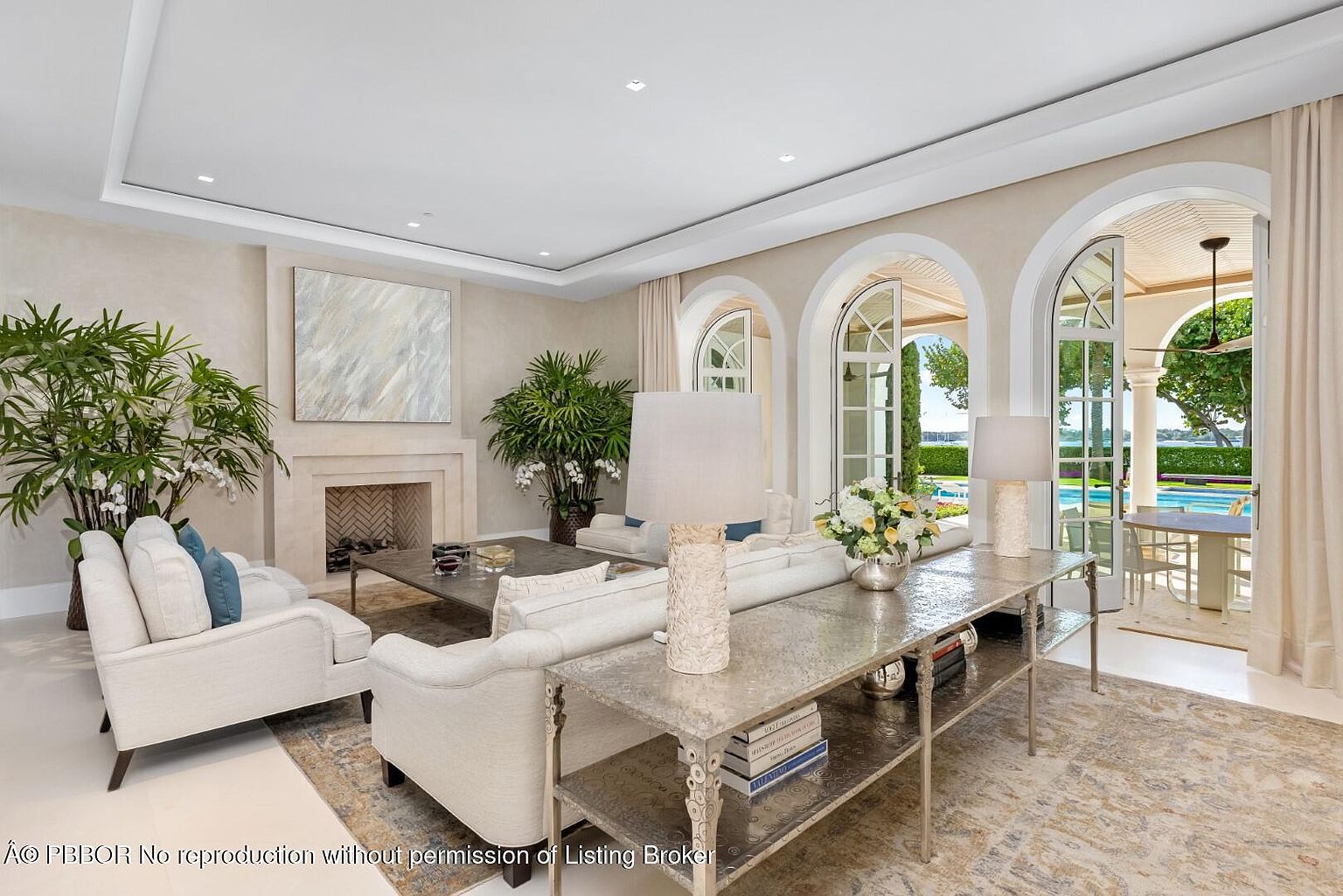 This is an interior shot of a luxurious living room featuring elegant furnishings and architectural details. The room is bathed in natural light streaming through arched doorways that lead to an outdoor area with a pool. The neutral color palette, combined with the plush seating and decorative plants, creates a sophisticated and inviting atmosphere.
