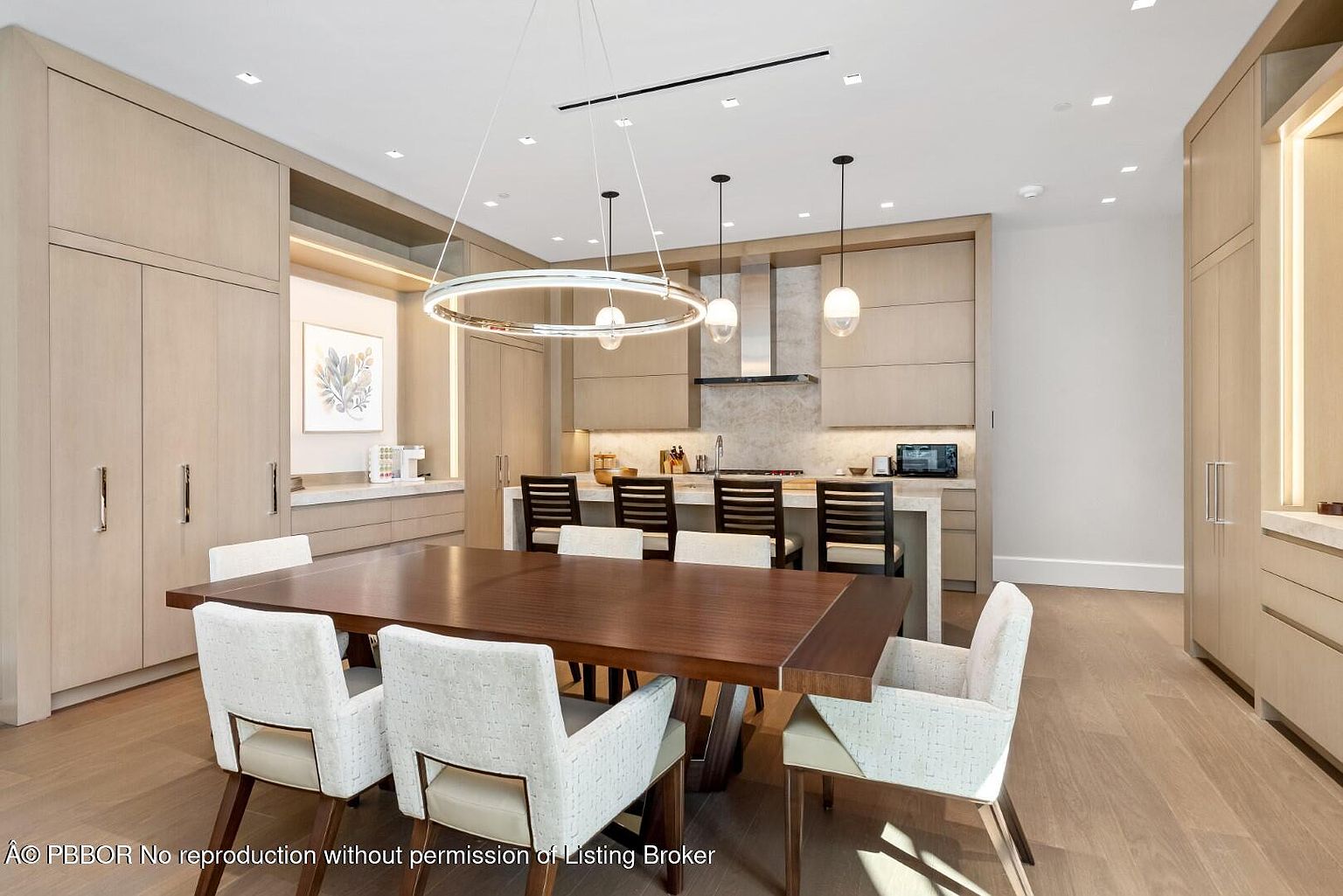 This interior shot showcases a dining area adjacent to a modern kitchen. The dining room features a large wooden table surrounded by upholstered chairs, illuminated by a circular chandelier. The kitchen area includes sleek cabinetry, a kitchen island with bar stools, and modern appliances, creating a cohesive and stylish living space.