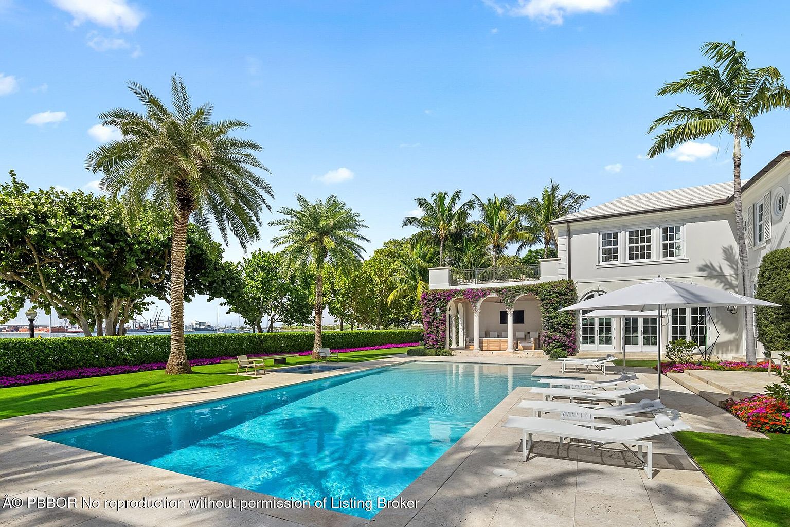 This image showcases a luxurious backyard featuring a large rectangular swimming pool with crystal clear water, surrounded by a meticulously manicured lawn and lush landscaping. Several lounge chairs are arranged on the stone patio beside the pool, and a grand white house with a covered patio adorned with flowering vines stands in the background, creating an inviting and serene outdoor living space.