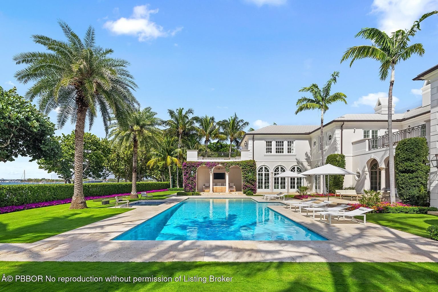 This image showcases a luxurious backyard featuring a large rectangular pool with crystal clear blue water, surrounded by a travertine patio and lush green lawn. The property includes a grand white house with arched windows and a covered outdoor living area adorned with purple flowers, complemented by tall palm trees and manicured hedges, creating a serene and upscale atmosphere.