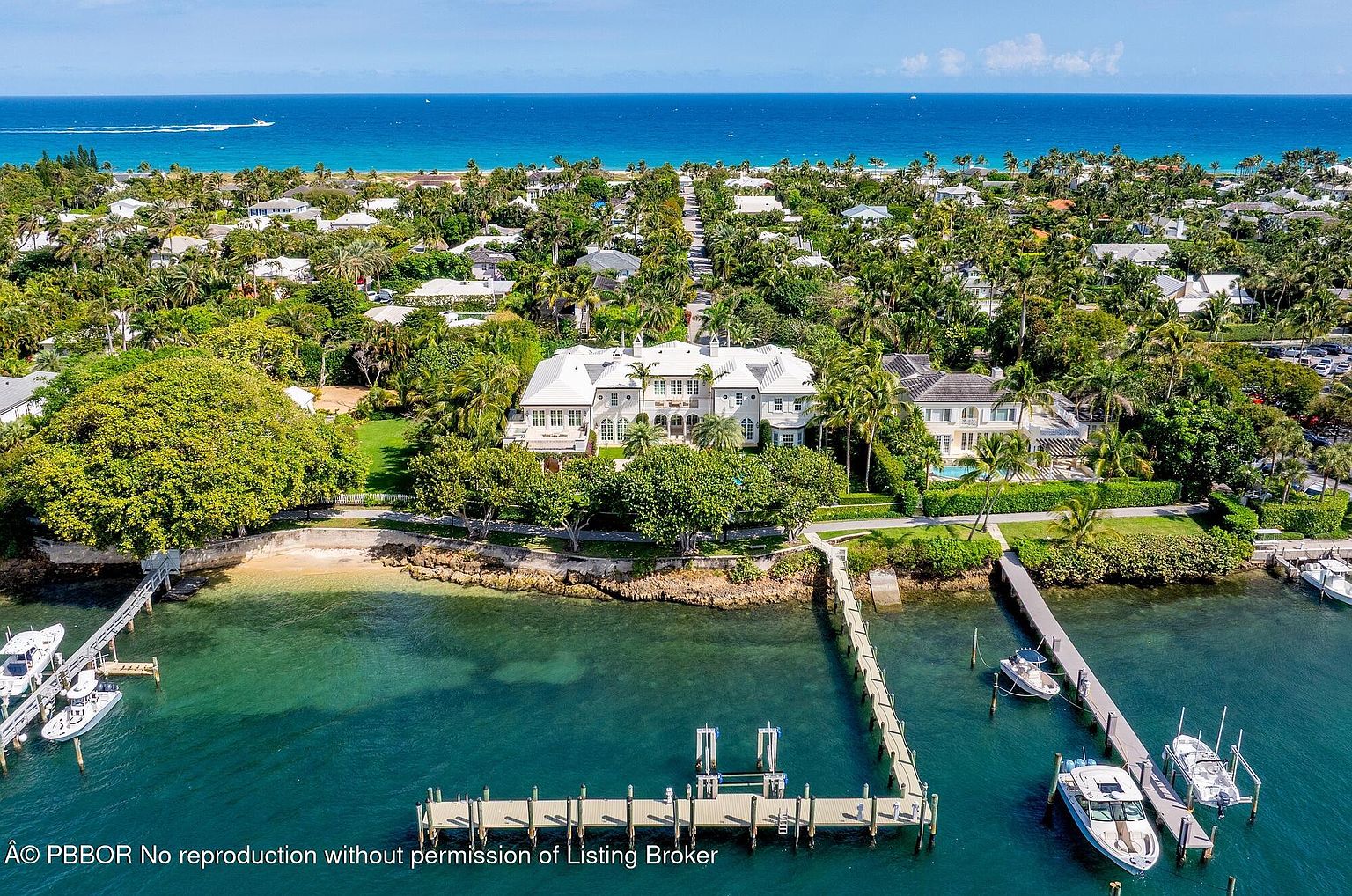 This aerial view showcases a luxurious waterfront estate with a private dock and multiple boats. The property features a large, elegant house surrounded by lush landscaping and mature trees. The turquoise water and clear blue sky create a stunning backdrop, emphasizing the property's prime location and upscale appeal.