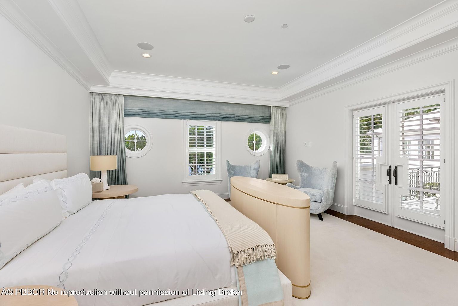 This is a bright and airy primary bedroom featuring a large bed with white linens, complemented by light blue accents. The room has a sophisticated design with white walls, crown molding, and round windows, creating a serene and luxurious atmosphere. French doors provide access to an outdoor space, enhancing the room's appeal.