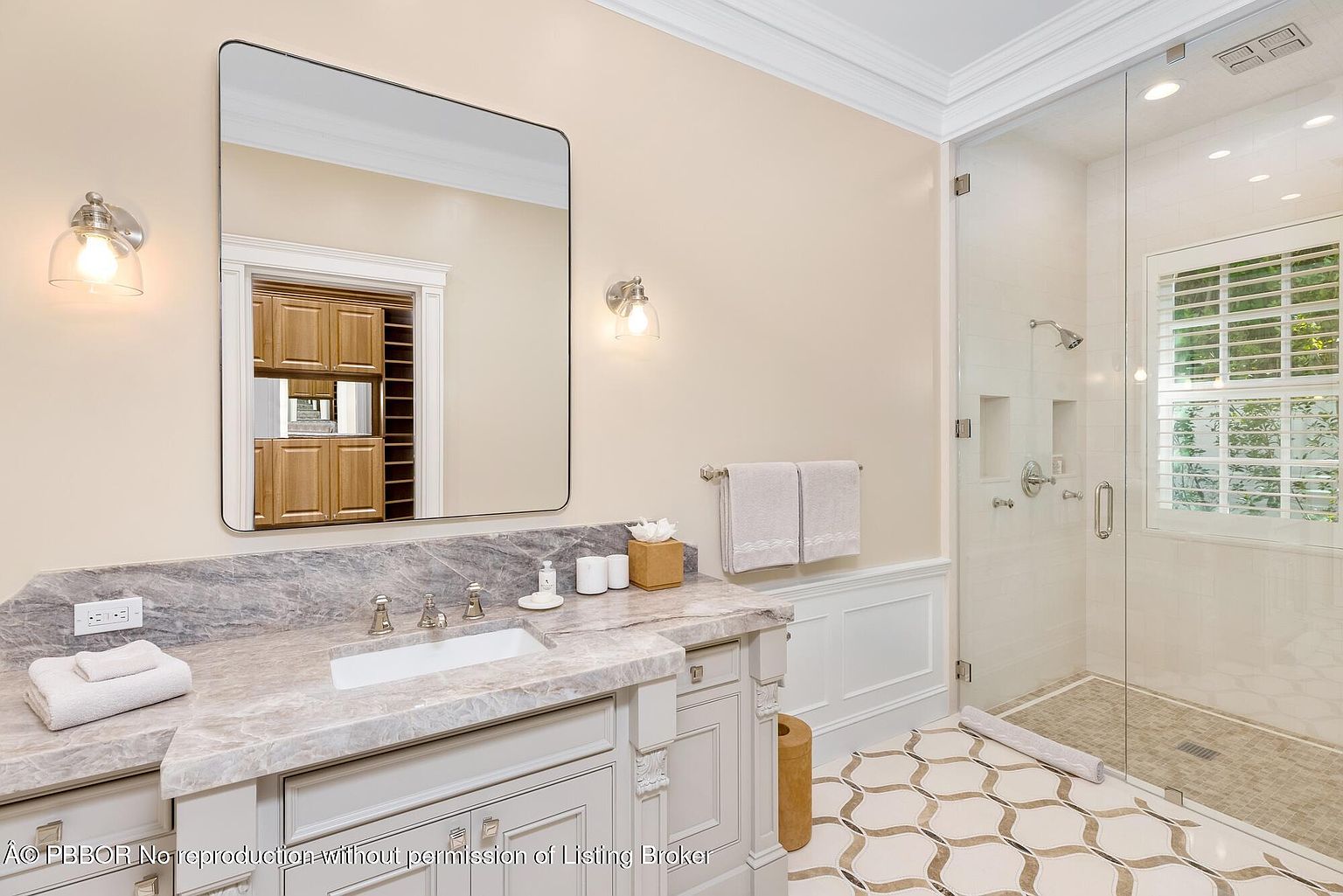 This is a well-lit primary bathroom featuring a marble countertop vanity with a white sink and chrome fixtures. A large mirror reflects a glimpse into an adjacent room, and sconces provide soft lighting. The shower is enclosed in glass, showcasing a tiled interior, and the floor is decorated with a patterned tile design.
