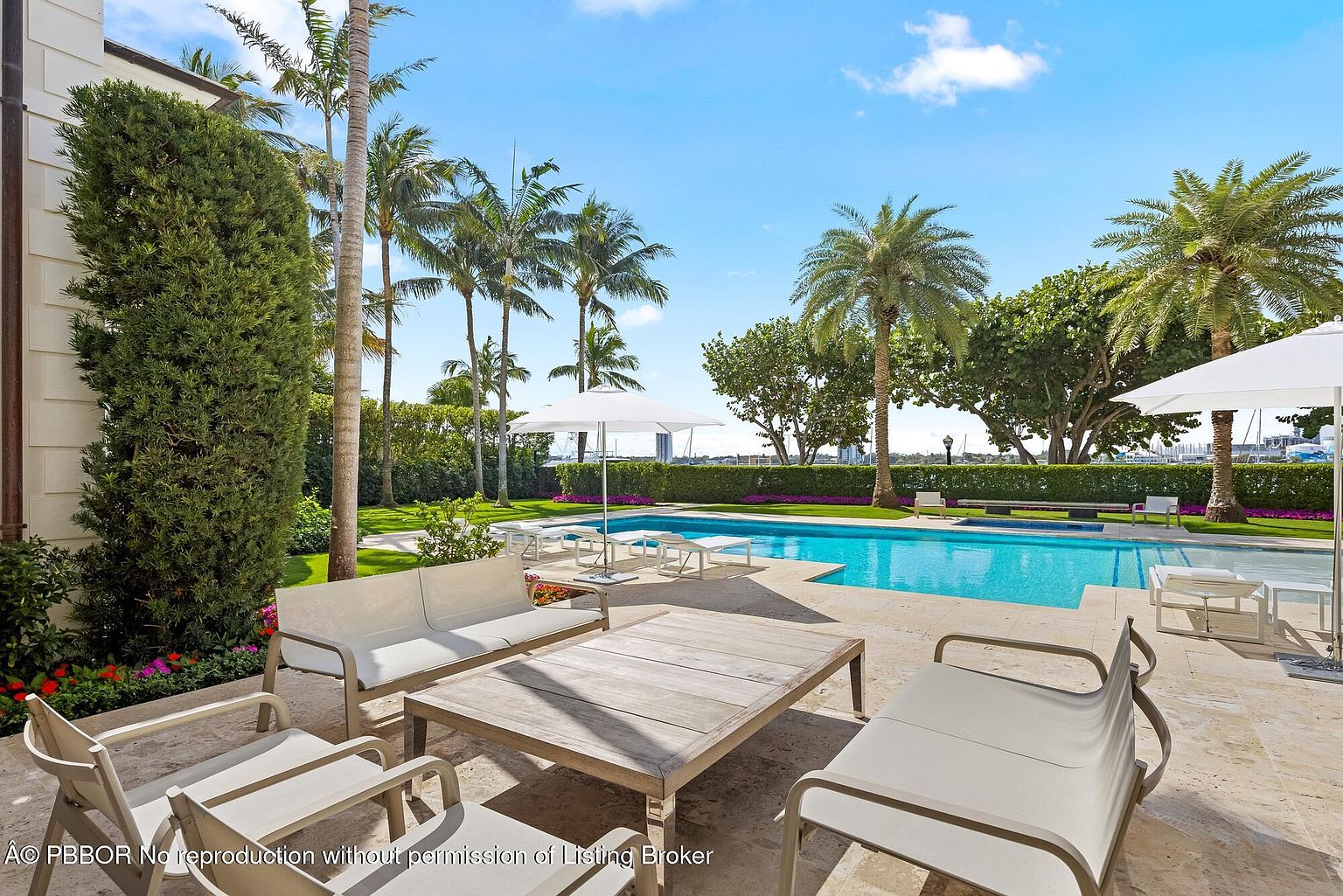 This image showcases a luxurious outdoor pool area, complete with modern patio furniture, including a sofa, chairs, and a coffee table. The pool is surrounded by lush landscaping, including palm trees and manicured hedges, creating a private and serene atmosphere. The view extends to a waterfront area with boats, suggesting a high-end property with direct water access.