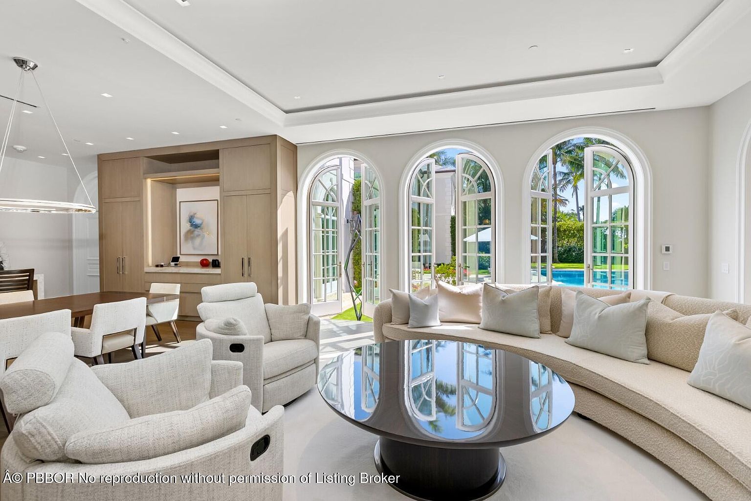 This is a bright and airy living room featuring a curved sofa with plush pillows, two comfortable armchairs, and a reflective round coffee table. The room is illuminated by natural light streaming through arched windows that offer views of the outdoor pool and landscaping. The overall aesthetic is luxurious and inviting, perfect for relaxation and entertaining.