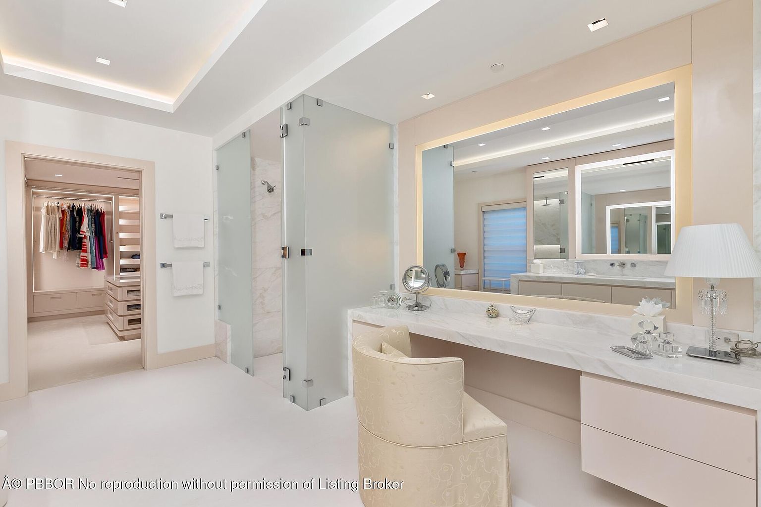 This is a bright and luxurious primary bathroom featuring a vanity area with a large mirror and a comfortable chair. A glass-enclosed shower is visible in the background, and a walk-in closet is accessible through a doorway on the left. The color palette is soft and neutral, creating a serene and elegant atmosphere.