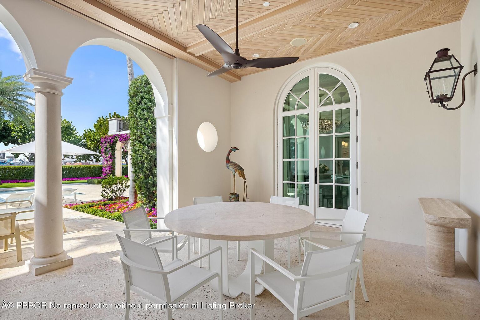 This image showcases a covered patio area, featuring a round dining table with white chairs, set against a backdrop of manicured gardens visible through arched openings. The ceiling has a decorative wood pattern, and a ceiling fan adds to the comfort of the space. An elegant outdoor lantern is mounted on the wall, and a decorative bird statue adds a touch of sophistication.
