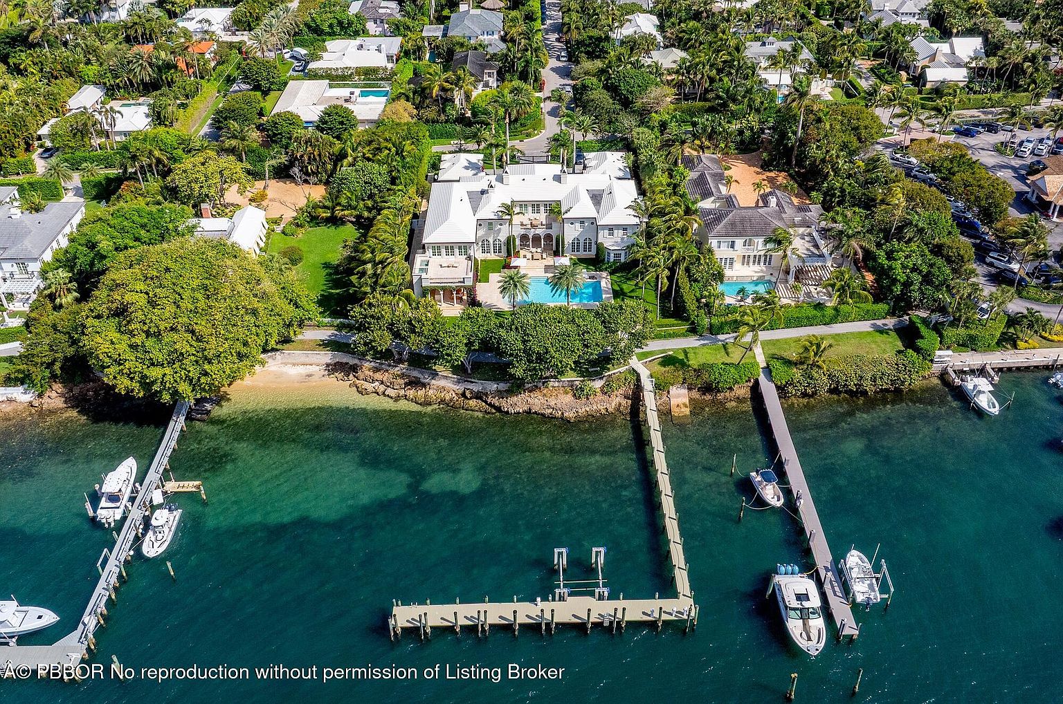 This aerial shot showcases a luxurious waterfront property featuring a grand mansion with a pool and meticulously landscaped grounds. The property includes private docks with boats, emphasizing its exclusive waterfront access and upscale lifestyle. Lush greenery surrounds the estate, providing privacy and enhancing the overall appeal.