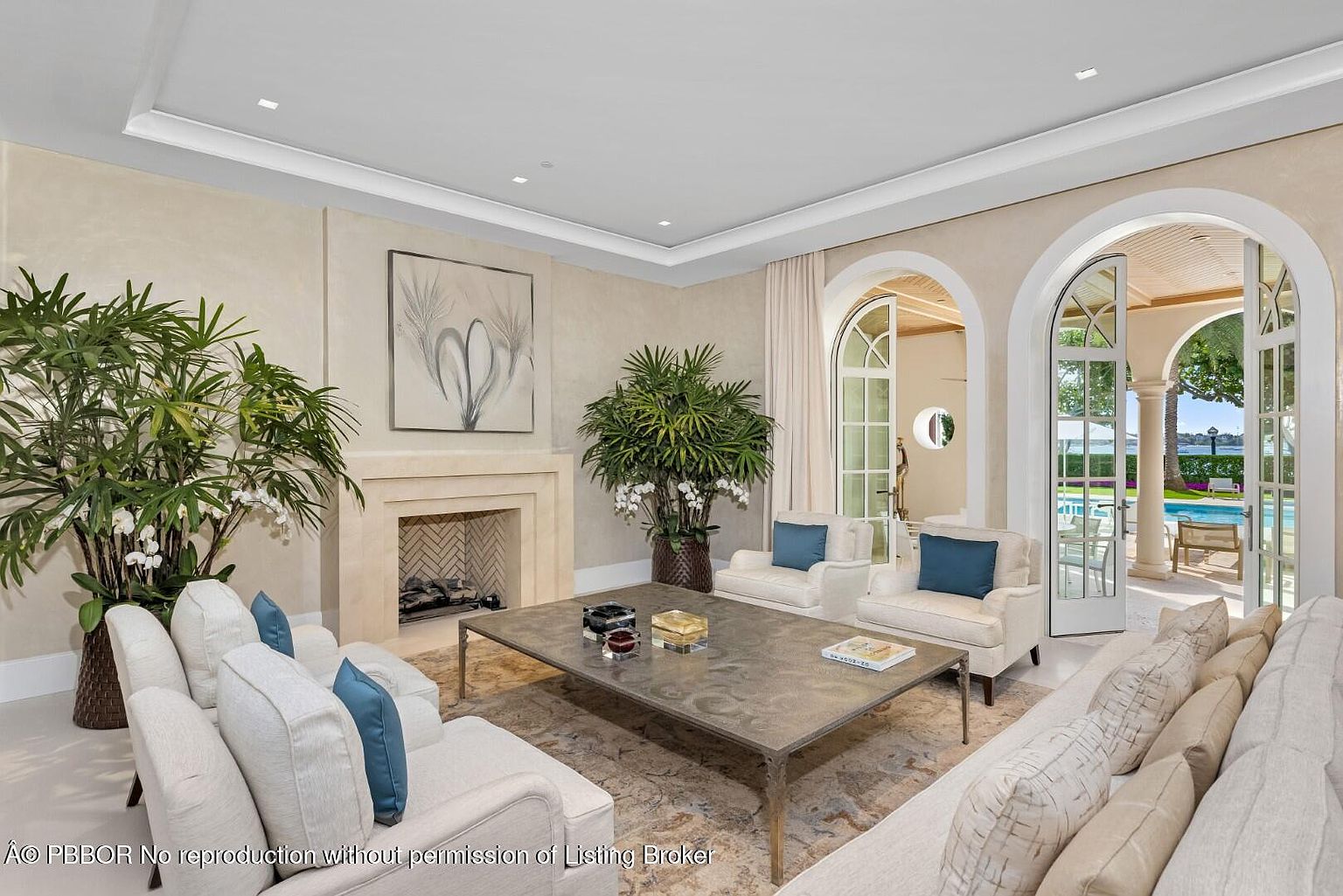 This is an interior shot of a luxurious living room featuring elegant furnishings and decor. The room is bathed in natural light streaming through arched doorways that lead to an outdoor pool area. The neutral color palette, combined with the plush seating and sophisticated artwork, creates a serene and inviting atmosphere.