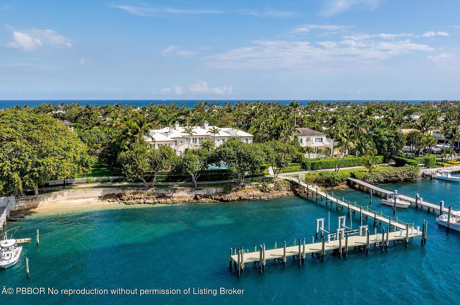 This aerial view showcases a luxurious waterfront estate with a pristine white mansion nestled among lush greenery and palm trees. The property features a private beach, a long pier extending into the turquoise water, and well-manicured lawns, creating an impression of exclusivity and serene coastal living. The overall composition highlights the property's prime location and upscale amenities.