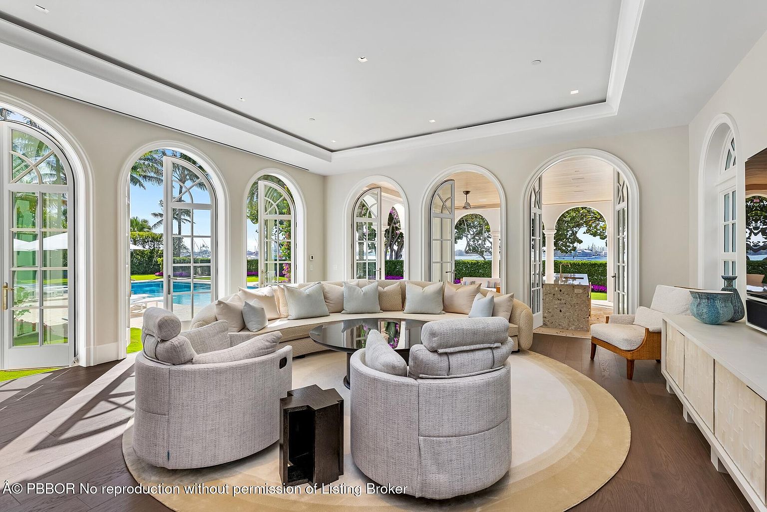 This is a bright and airy living room featuring a curved sofa, two armchairs, and a round coffee table on a circular rug. The room is illuminated by natural light streaming through arched windows and doors, offering views of the outdoor pool and landscaping. The overall aesthetic is luxurious and inviting, perfect for relaxation and entertaining.