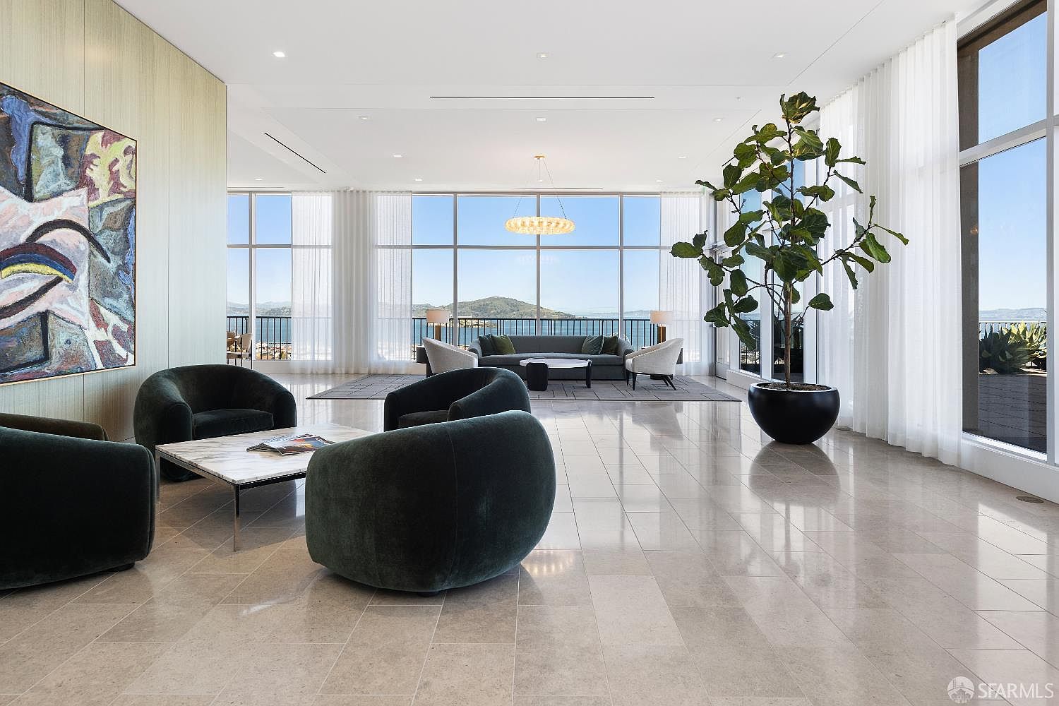 This expansive, modern living area features a minimalist aesthetic with polished stone flooring and a neutral color palette. The room is anchored by several dark, curved velvet armchairs and a central seating group, all positioned to take advantage of the panoramic water and mountain views framed by floor-to-ceiling windows. A large, abstract painting adds a vibrant focal point to the left wall, while a tall potted fiddle-leaf fig tree introduces a natural element to the bright, airy space.