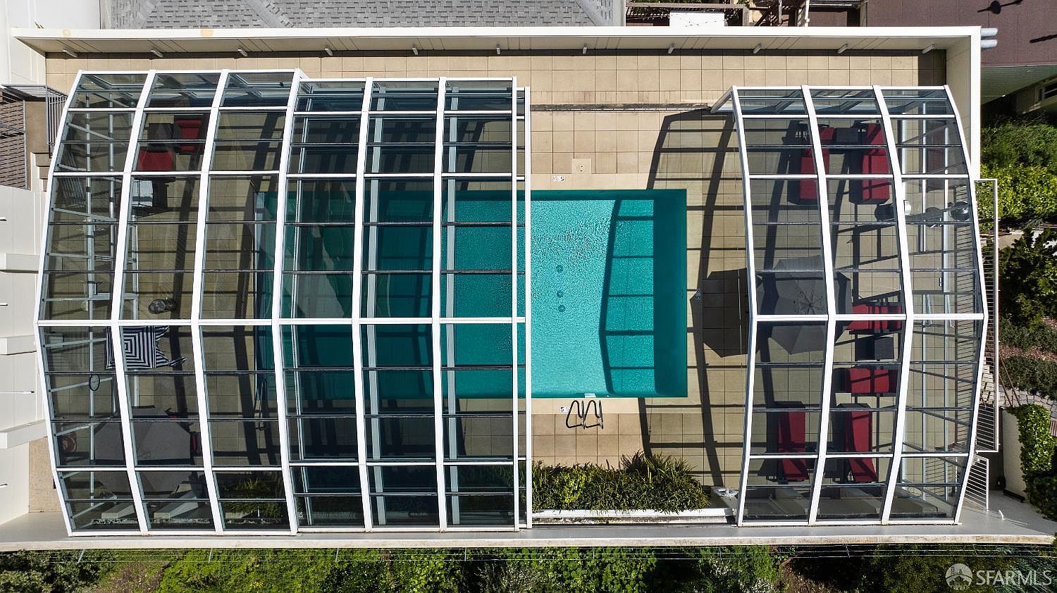 This top-down aerial view captures a rectangular swimming pool enclosed within a large, retractable glass-paneled structure. The pool deck is paved with neutral-toned tiles and features several lounge chairs and a patio umbrella, creating a private and sheltered recreational space. The perspective emphasizes the symmetry of the enclosure and the clear, blue water of the pool, highlighting a well-maintained outdoor amenity.