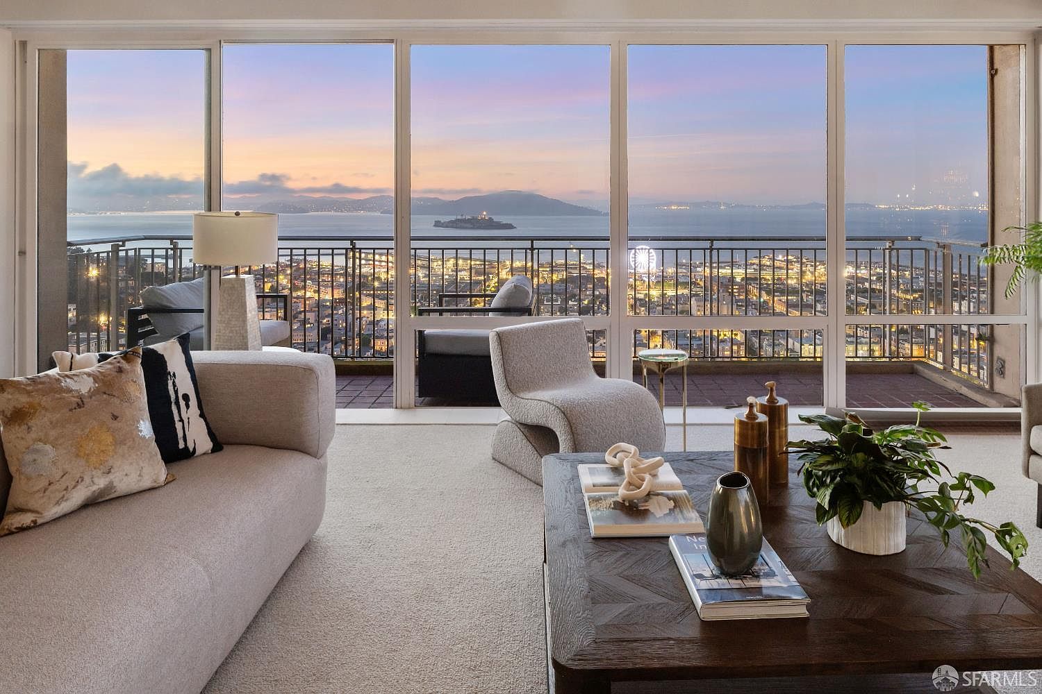 This sophisticated living room features a neutral color palette and a contemporary aesthetic, anchored by a plush sofa and a sculptural accent chair. The space is defined by expansive floor-to-ceiling windows that frame a breathtaking panoramic view of the bay, Alcatraz Island, and the city skyline at dusk. A dark wood coffee table with decorative objects and a potted plant adds a touch of warmth to the bright, airy interior.