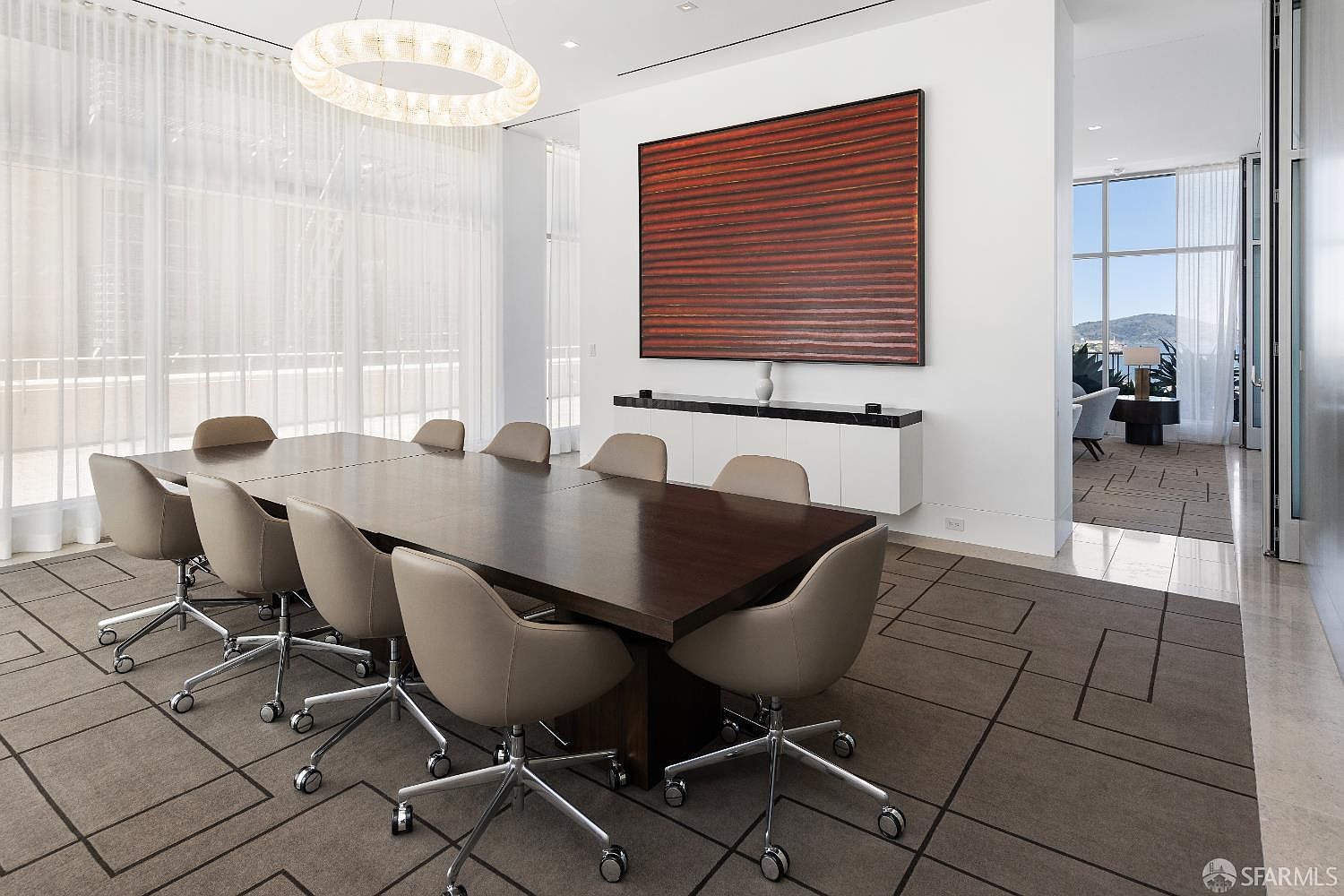 This sophisticated dining room features a large, dark-toned rectangular table surrounded by ten modern, light-colored upholstered chairs on casters. A striking, circular tiered chandelier hangs above the table, while a long, minimalist white sideboard with a dark top sits against the wall beneath a large, textured red abstract painting. The room is defined by a unique geometric-patterned carpet and expansive floor-to-ceiling windows that allow for abundant natural light and a view into an adjacent living area.