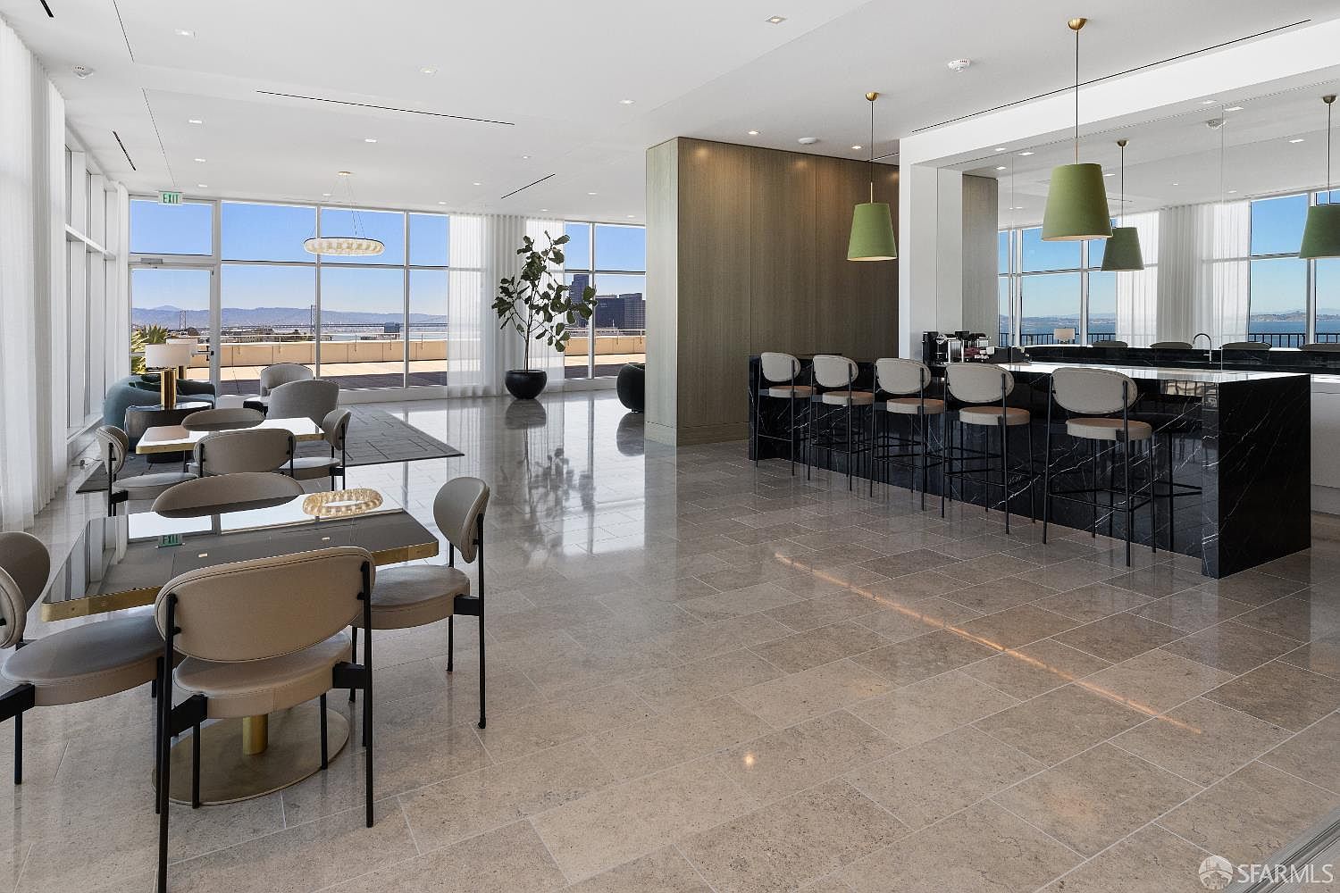 This sophisticated lounge area features a sleek black stone bar with barstool seating and a separate dining area with modern chairs and tables. The space is defined by polished stone flooring and large windows that offer expansive city and bay views. The overall aesthetic is contemporary and upscale, designed for social gatherings or professional use.