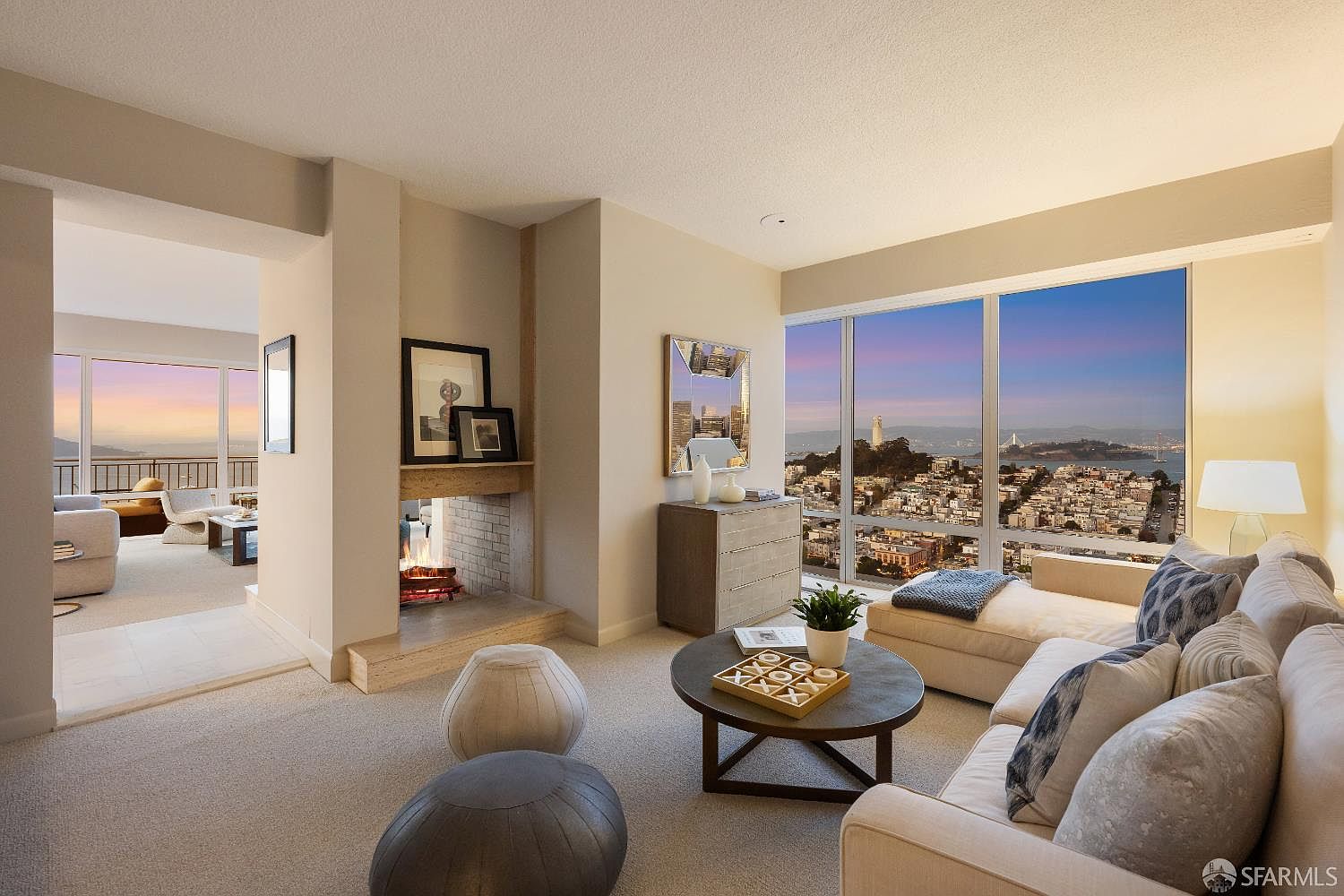 This sophisticated living room features a neutral color palette and a comfortable seating area centered around a round coffee table. A double-sided fireplace with a stone-clad chimney breast serves as a focal point, connecting the main living space to an adjacent room with a balcony. Large windows provide expansive, panoramic views of the city skyline and the bay, creating a bright and airy atmosphere.