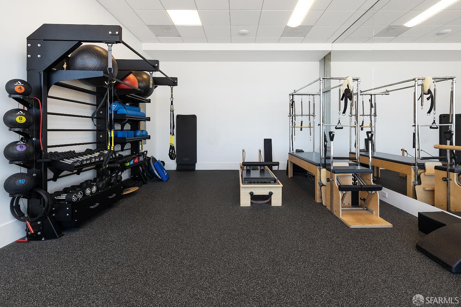 This well-equipped home gym features a dedicated storage rack stocked with medicine balls, dumbbells, and various fitness accessories. To the right, a professional-grade Pilates reformer sits on the dark, textured flooring, with a large mirror reflecting the equipment to create a sense of depth. The space is brightly lit by recessed ceiling lights, offering a clean and functional environment for exercise.