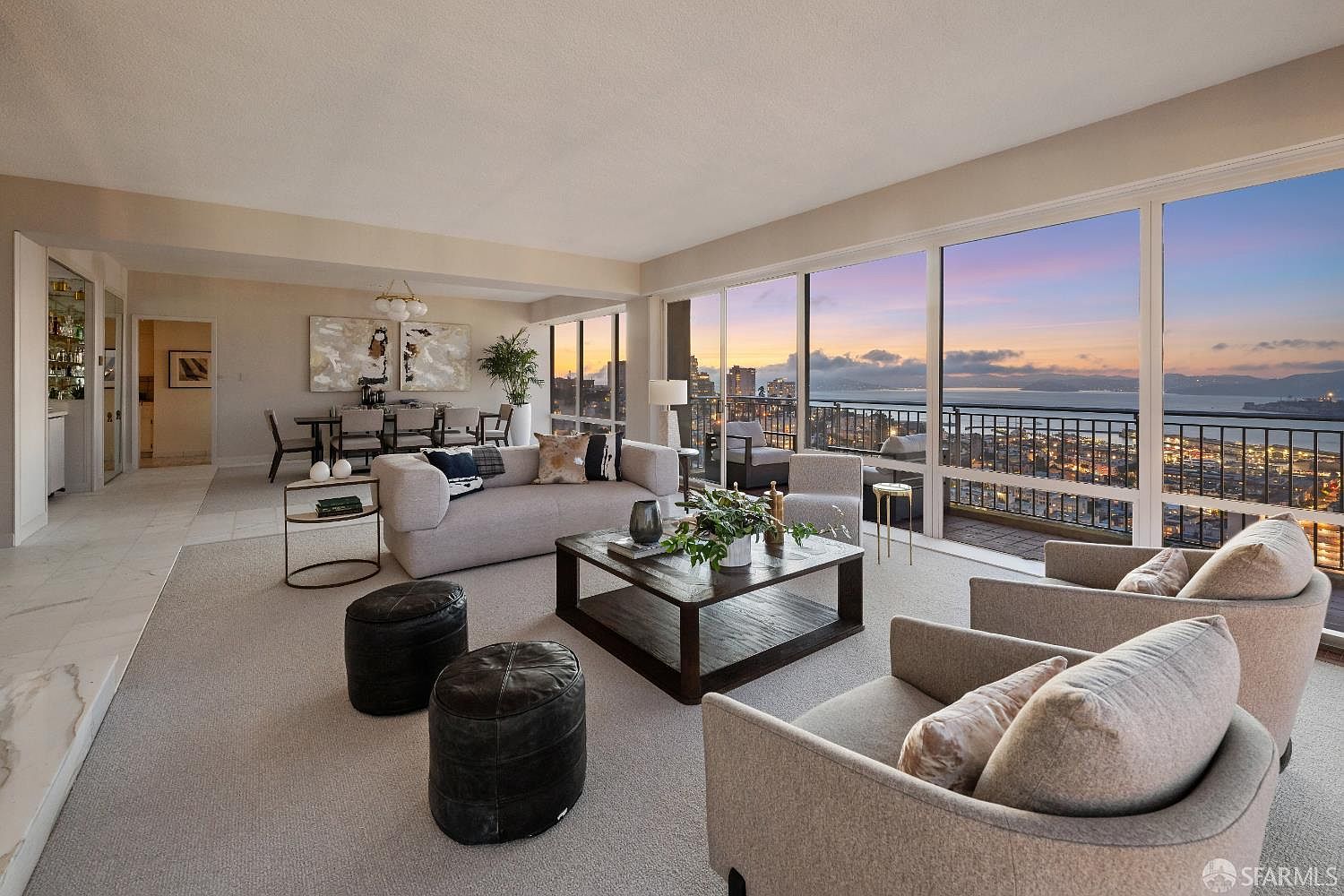 This sophisticated living room features an open-concept layout with a neutral color palette and plush, contemporary furnishings. Large, expansive windows provide a panoramic view of the city skyline and bay at sunset, bathing the space in warm, natural light. The room is anchored by a dark wood coffee table and two leather ottomans, creating a refined yet comfortable atmosphere perfect for entertaining.