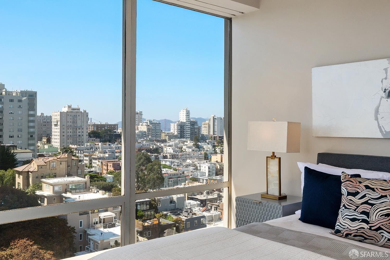 This bedroom features expansive floor-to-ceiling windows that frame a panoramic urban cityscape, offering a bright and airy atmosphere. The room is furnished with a bed dressed in neutral-toned linens, accented by a patterned throw pillow, and a bedside table topped with a modern lamp featuring a rectangular shade. The perspective captures the contrast between the serene, minimalist interior and the bustling city view outside.