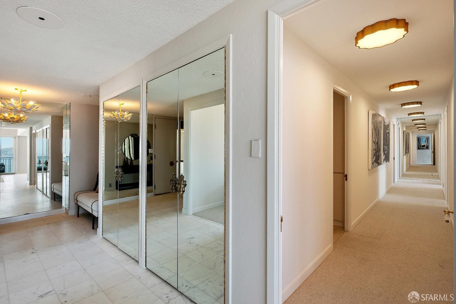 This interior view captures a bright, expansive entryway and hallway featuring light-colored stone flooring that transitions into plush carpeting. The space is defined by large mirrored closet doors that reflect the surrounding area, creating an open and airy atmosphere, while a long corridor extends into the distance, illuminated by a series of flush-mount ceiling fixtures. The perspective provides a clear transition from the tiled foyer into the carpeted hallway, highlighting the clean, neutral aesthetic of the home.