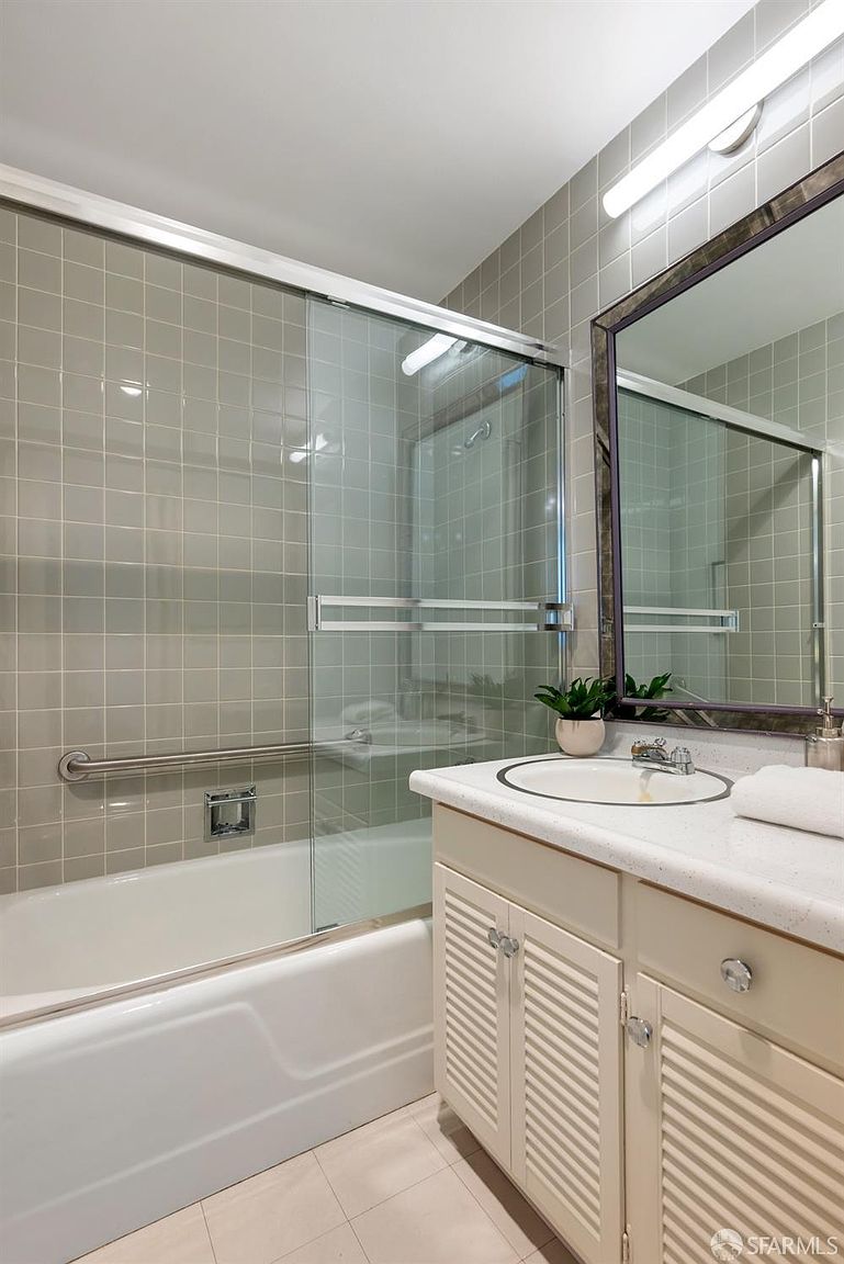 This bathroom features a neutral color palette with light-toned, louvered-style vanity cabinetry topped with a solid-surface counter and an integrated sink. A sliding glass shower door encloses a tub area lined with square tiles and equipped with a horizontal grab bar for accessibility. The space is illuminated by a long, linear light fixture mounted above a framed mirror, creating a clean and functional aesthetic.