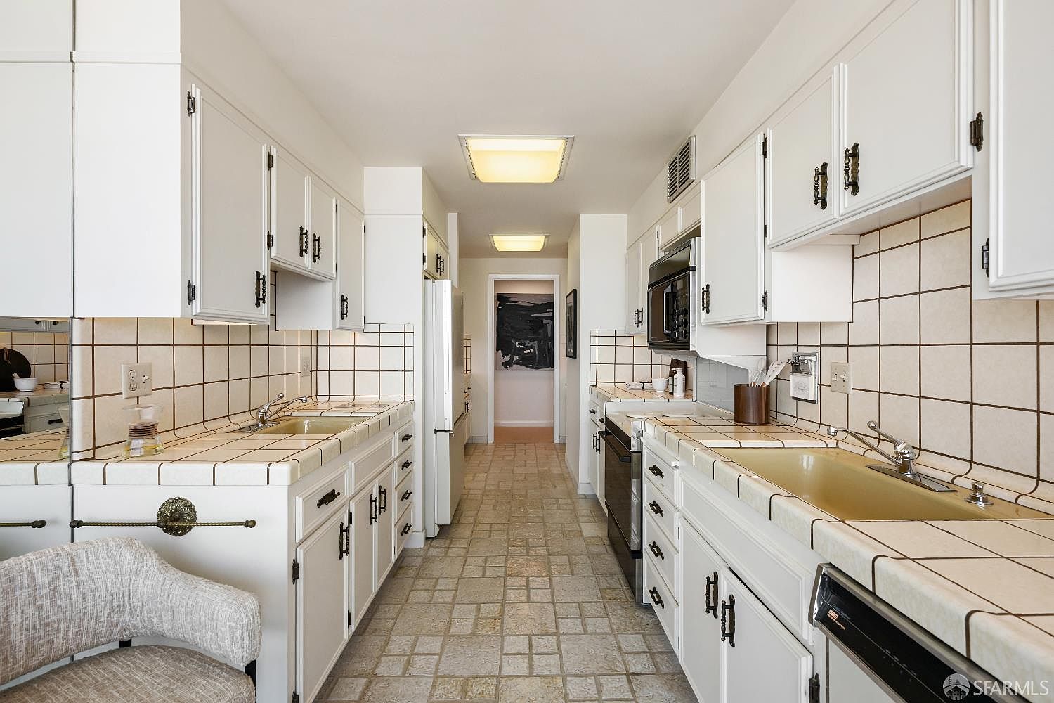 This galley-style kitchen features white cabinetry with dark hardware and tiled countertops that extend into a backsplash. The space is illuminated by two flush-mount ceiling lights and includes a double sink, a built-in microwave, and a dishwasher. A neutral-toned tiled floor runs throughout the kitchen, leading toward a doorway that opens into another room.