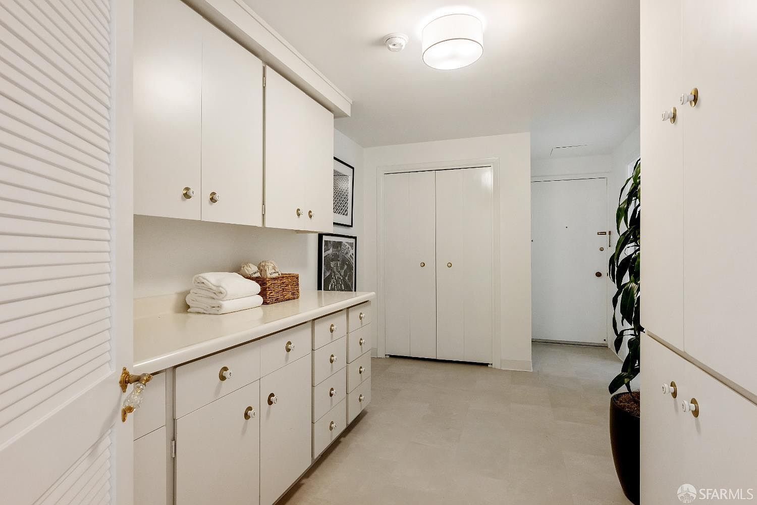 This bright, neutral-toned hallway features extensive built-in cabinetry providing ample storage space. The area includes a long counter surface, a set of double doors leading to a closet, and a doorway at the end of the hall. A tall potted plant adds a touch of greenery to the clean, minimalist aesthetic.