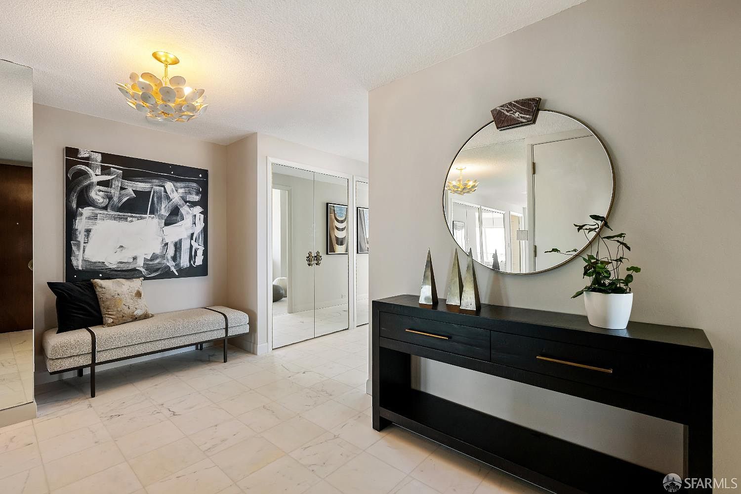 This elegant entryway features light-colored stone flooring and a sophisticated, modern aesthetic. A long bench with a patterned cushion sits beneath a bold, abstract black-and-white painting, while a sleek black console table with a circular mirror creates a refined focal point on the opposite wall. The space is illuminated by a unique, multi-petaled light fixture, and mirrored closet doors add depth and brightness to the hallway.