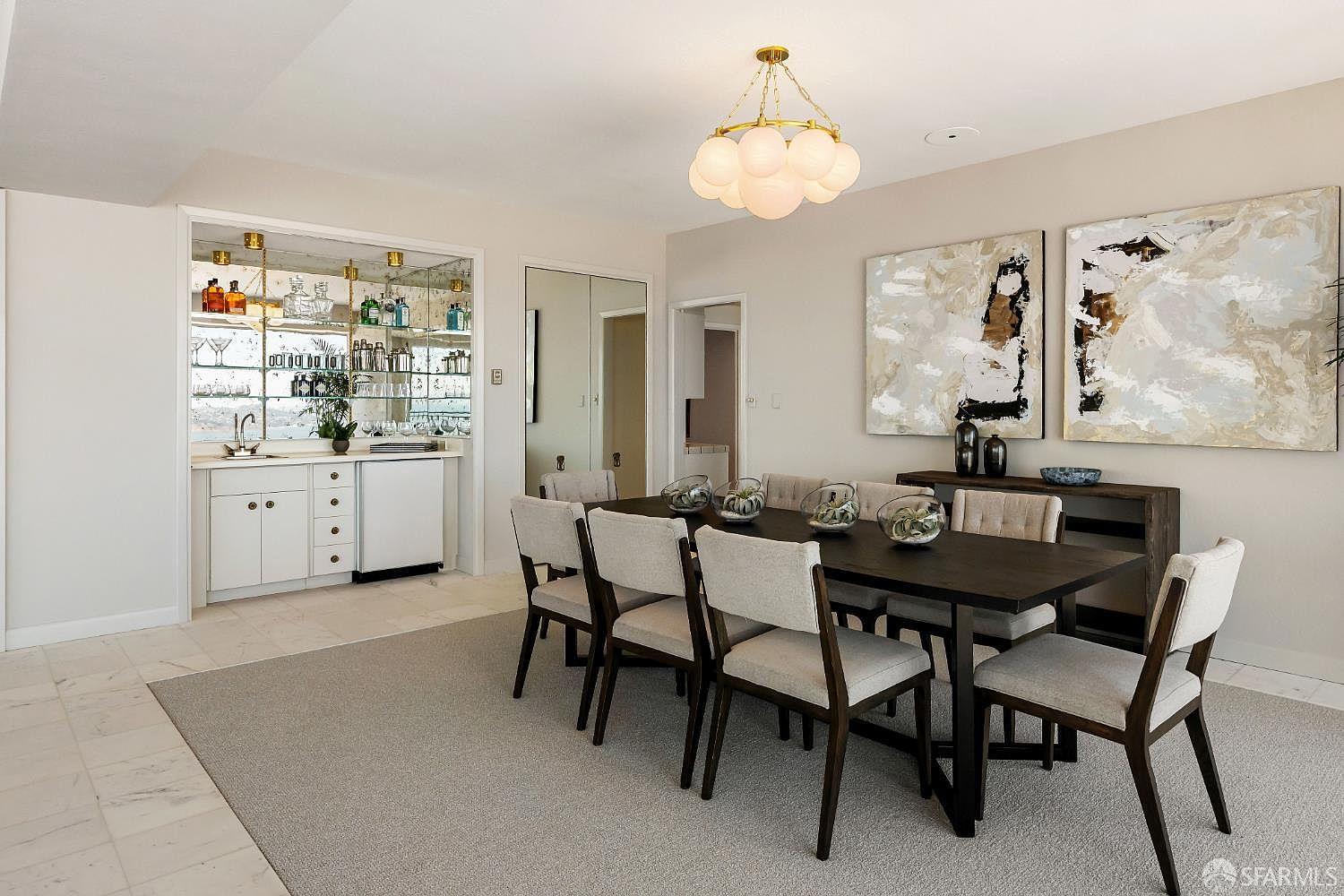 This elegant dining room features a dark wood table surrounded by six upholstered chairs, set atop a neutral area rug. A modern, multi-globe chandelier hangs above the table, while a mirrored wet bar with glass shelving and cabinetry provides a sophisticated touch to the space. The room is accented by two large abstract art pieces on the wall, creating a refined and cohesive atmosphere.