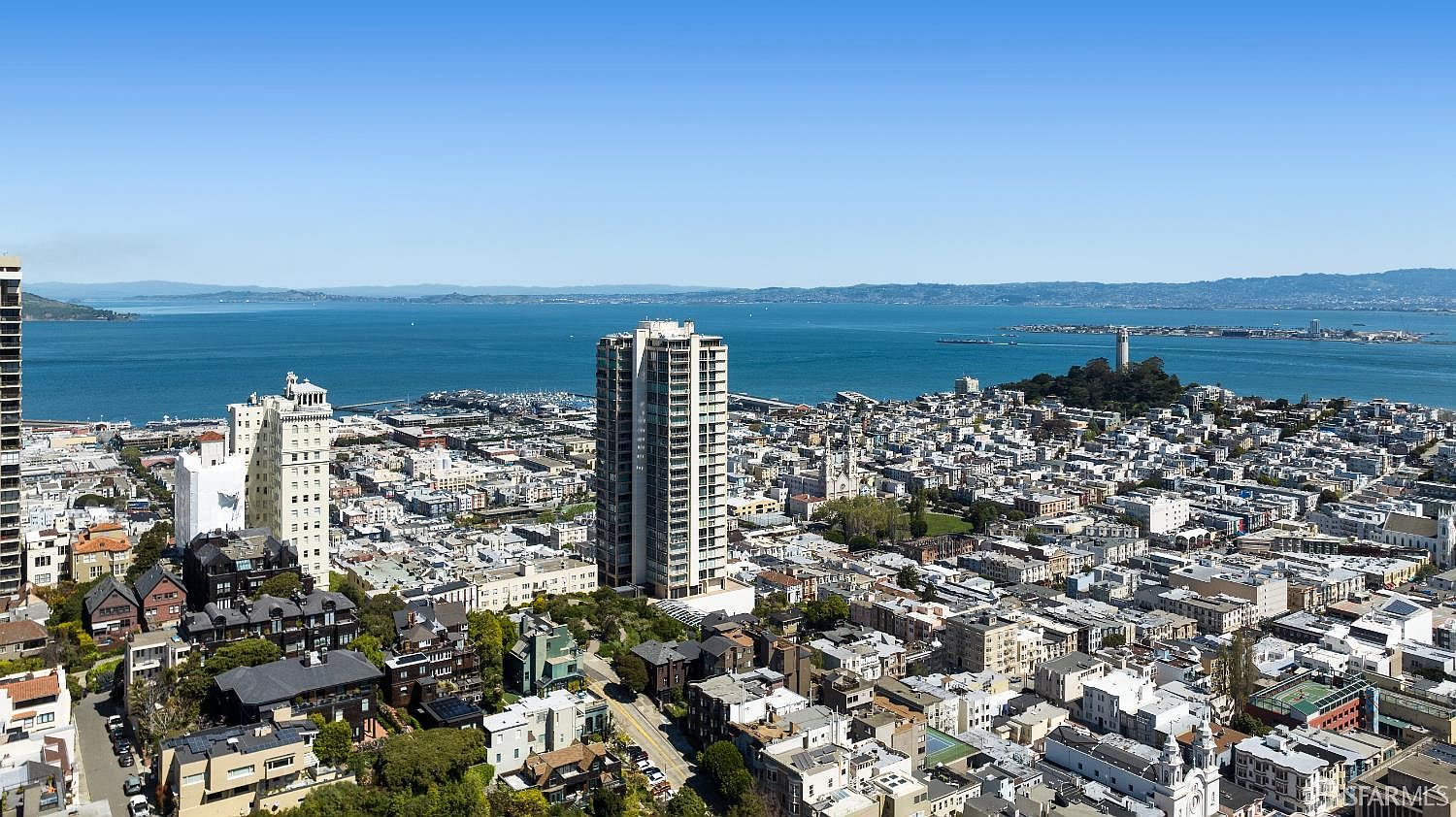 This high-angle aerial view captures a dense urban cityscape in San Francisco, showcasing a mix of historic residential architecture and modern high-rise buildings. The perspective looks out toward the expansive blue waters of the bay, with the iconic Coit Tower visible on the distant hillside. The scene provides a comprehensive look at the neighborhood's layout, density, and proximity to the waterfront.