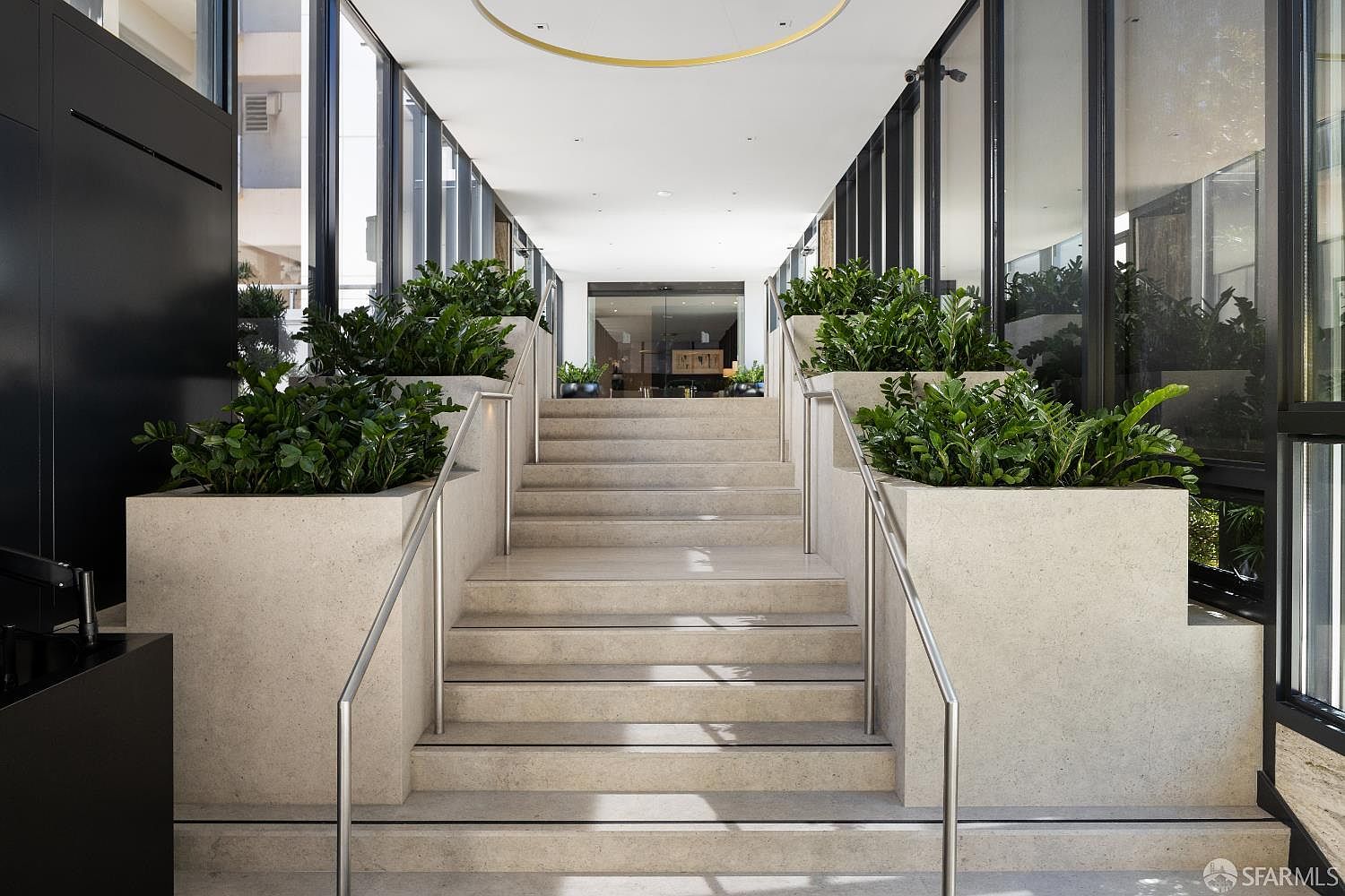 This elegant entryway features a wide, central staircase constructed from light-toned stone, flanked by integrated planters filled with lush greenery. The space is framed by tall glass panels on either side, creating a bright and airy atmosphere that leads toward a glass-doored entrance. A minimalist circular light fixture hangs from the ceiling, enhancing the modern, sophisticated aesthetic of this transition area.