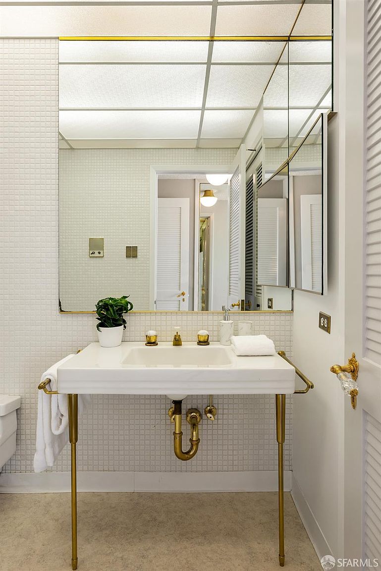This bathroom features a pedestal-style sink with gold-toned hardware and exposed plumbing, set against a wall of small white square tiles. A large, expansive mirror with a gold frame reflects the room, including a glimpse of a doorway and lighting fixtures. The space is bright and clean, offering a classic, vintage-inspired aesthetic.