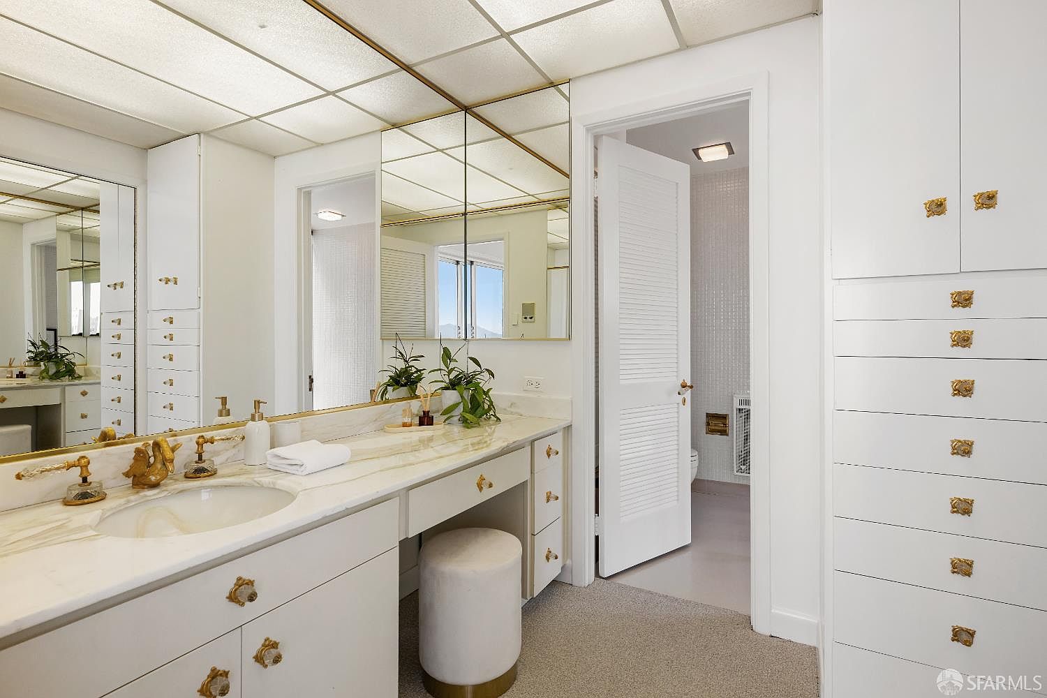 This primary bathroom features a long vanity with a stone countertop, integrated sink, and gold-toned hardware. A large, multi-pane mirror spans the wall above the vanity, reflecting the adjacent room and a doorway leading to a private water closet. The space is finished with white cabinetry, a neutral-toned stool, and a drop-ceiling with recessed lighting, creating a bright and classic aesthetic.