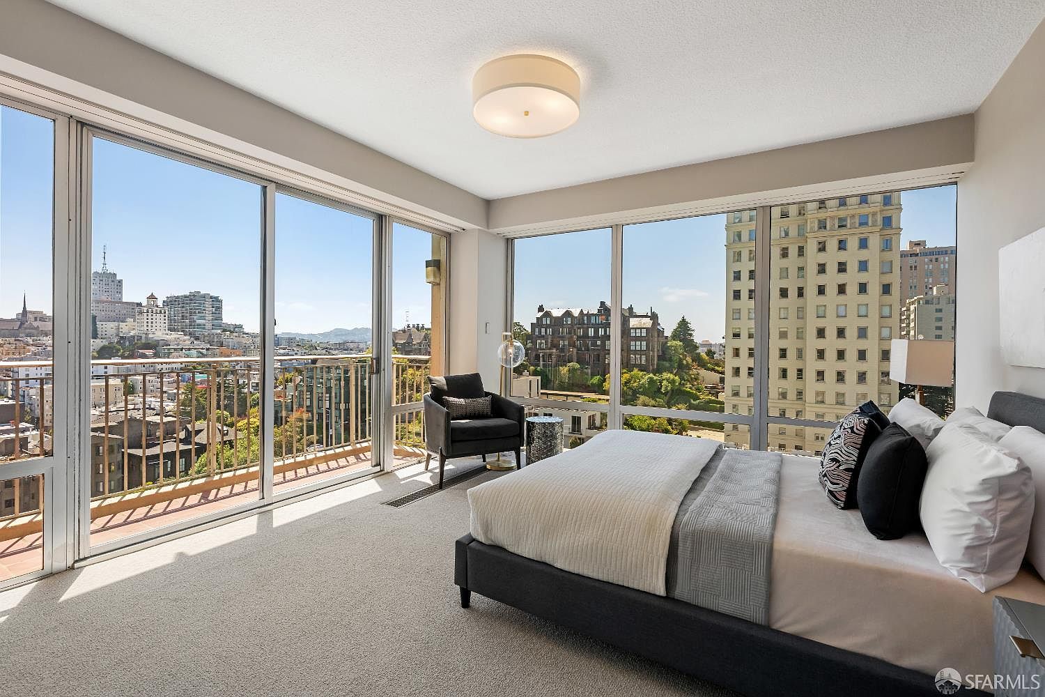 This primary bedroom features expansive floor-to-ceiling windows that offer panoramic views of the city skyline and surrounding architecture. The room is furnished with a modern bed dressed in neutral linens and a dark accent chair positioned near the glass. The space is bathed in natural light, creating an airy and sophisticated atmosphere.