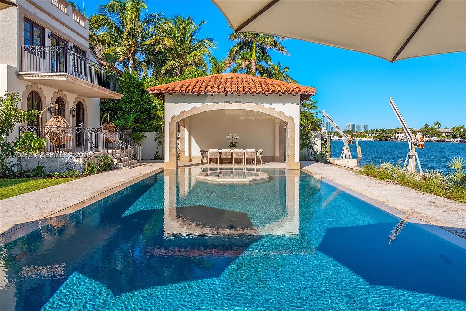 This luxurious waterfront backyard features a sparkling rectangular swimming pool with an integrated spa, set against a backdrop of a deep blue canal and distant city skyline. A covered, open-air pavilion with a terracotta-tiled roof provides a shaded dining area, while the surrounding stone patio offers ample space for relaxation. The scene is captured from a low-angle perspective, emphasizing the seamless transition between the elegant residence and the serene water views.