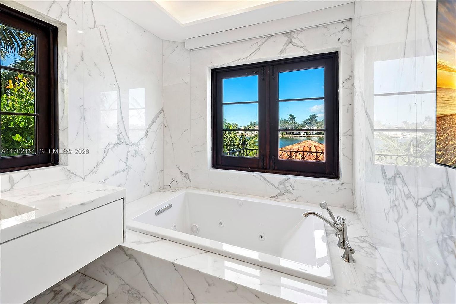 This luxurious primary bathroom features a deep soaking jetted tub encased in white stone with dramatic grey veining. The space is framed by dark-framed windows that offer a picturesque view of the water and tropical landscaping outside. The clean, bright aesthetic is enhanced by the reflective stone surfaces, creating a serene and high-end spa-like atmosphere.