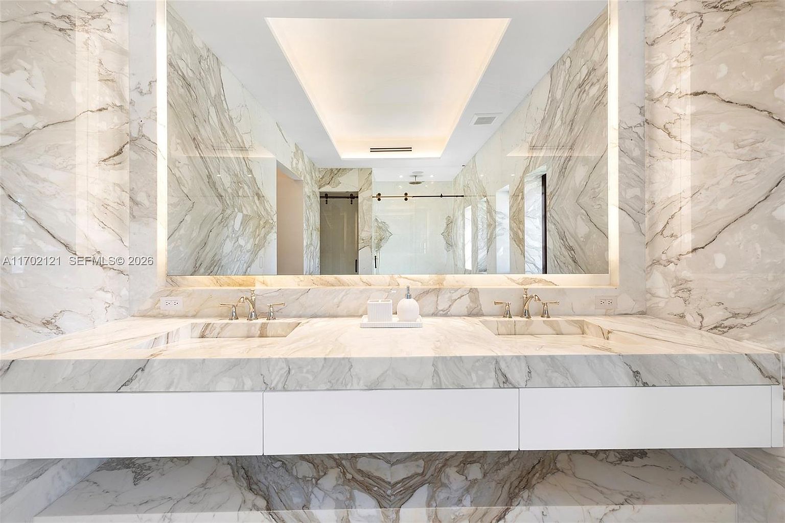 This luxurious primary bathroom features a double vanity with a continuous stone countertop and integrated sinks, set against a backdrop of dramatic, veined stone wall cladding. A massive, wall-to-wall mirror reflects the space, including a glass-enclosed shower area visible in the background, while integrated lighting provides a soft, ambient glow. The clean, minimalist design emphasizes high-end materials and a bright, sophisticated aesthetic.