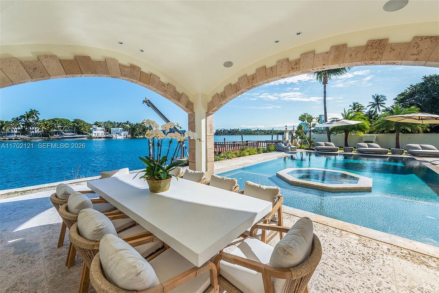 This luxurious covered patio features a large, light-toned dining table surrounded by woven chairs, perfect for outdoor entertaining. The space opens directly onto a pristine swimming pool and spa, with a stunning, unobstructed view of the sparkling blue bay beyond. The architectural design includes elegant stone arches that frame the waterfront scenery, creating a sophisticated and serene atmosphere.