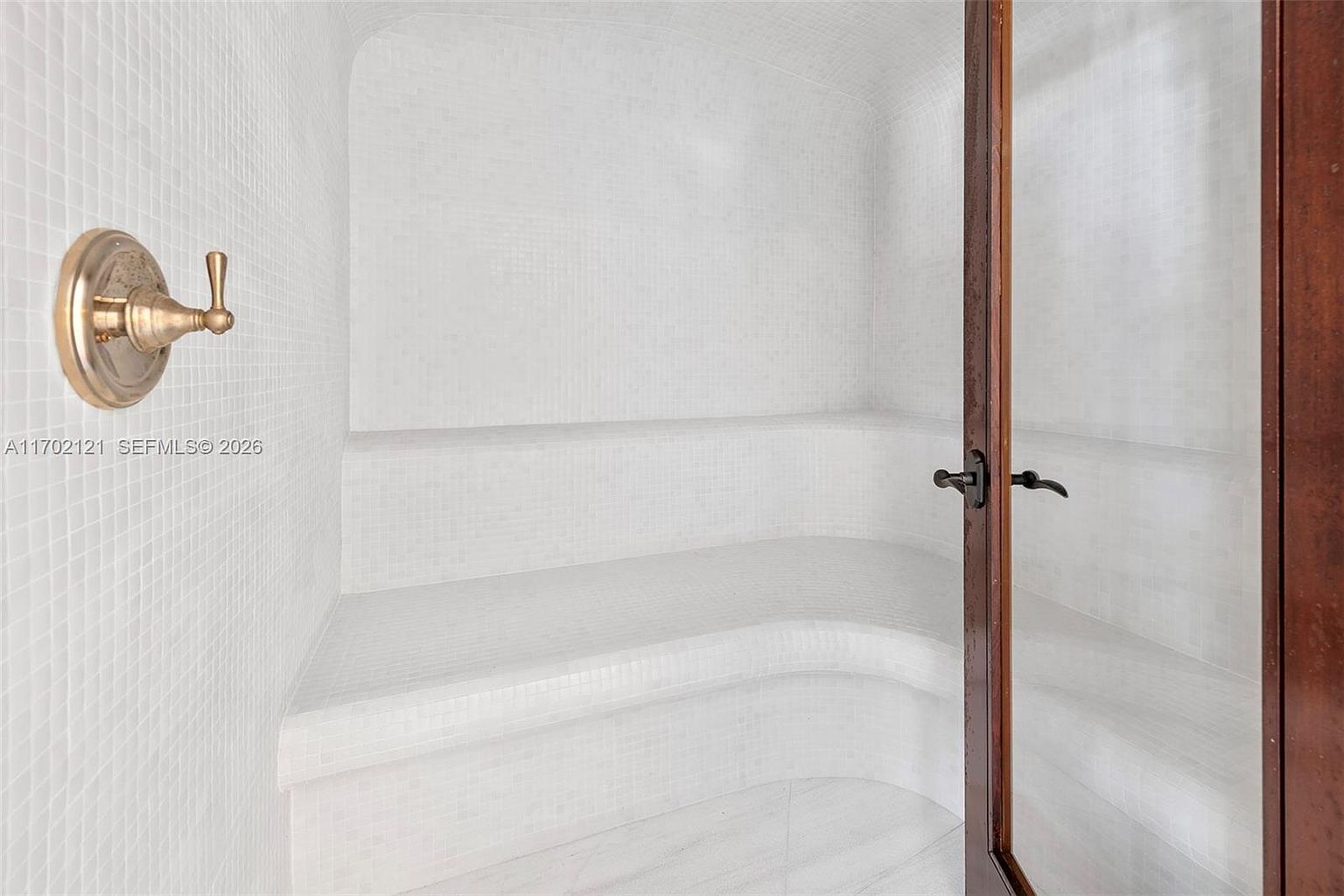 This image showcases a compact, tiled steam shower or sauna featuring built-in bench seating that follows the curved wall. The space is finished entirely in small, light-colored square tiles, creating a clean and minimalist aesthetic. A brass-toned fixture is mounted on the left wall, and a wooden-framed glass door provides entry to the enclosure.