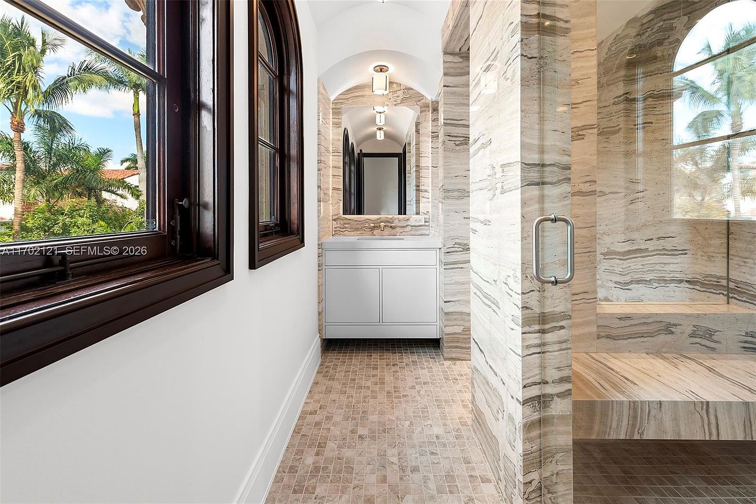 This luxurious bathroom features a sophisticated design with stone-clad walls and a walk-in shower enclosure. A white vanity with a sleek countertop sits beneath a mirror, framed by an arched architectural alcove. The space is brightened by natural light streaming through large windows that offer a view of palm trees outside.